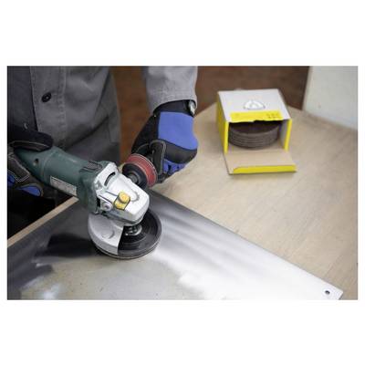 Eine Person benutzt einen Winkelschleifer an einem Metallblech und trägt Schutzhandschuhe. Eine Schachtel mit Trennscheiben liegt auf dem Tisch neben ihr.