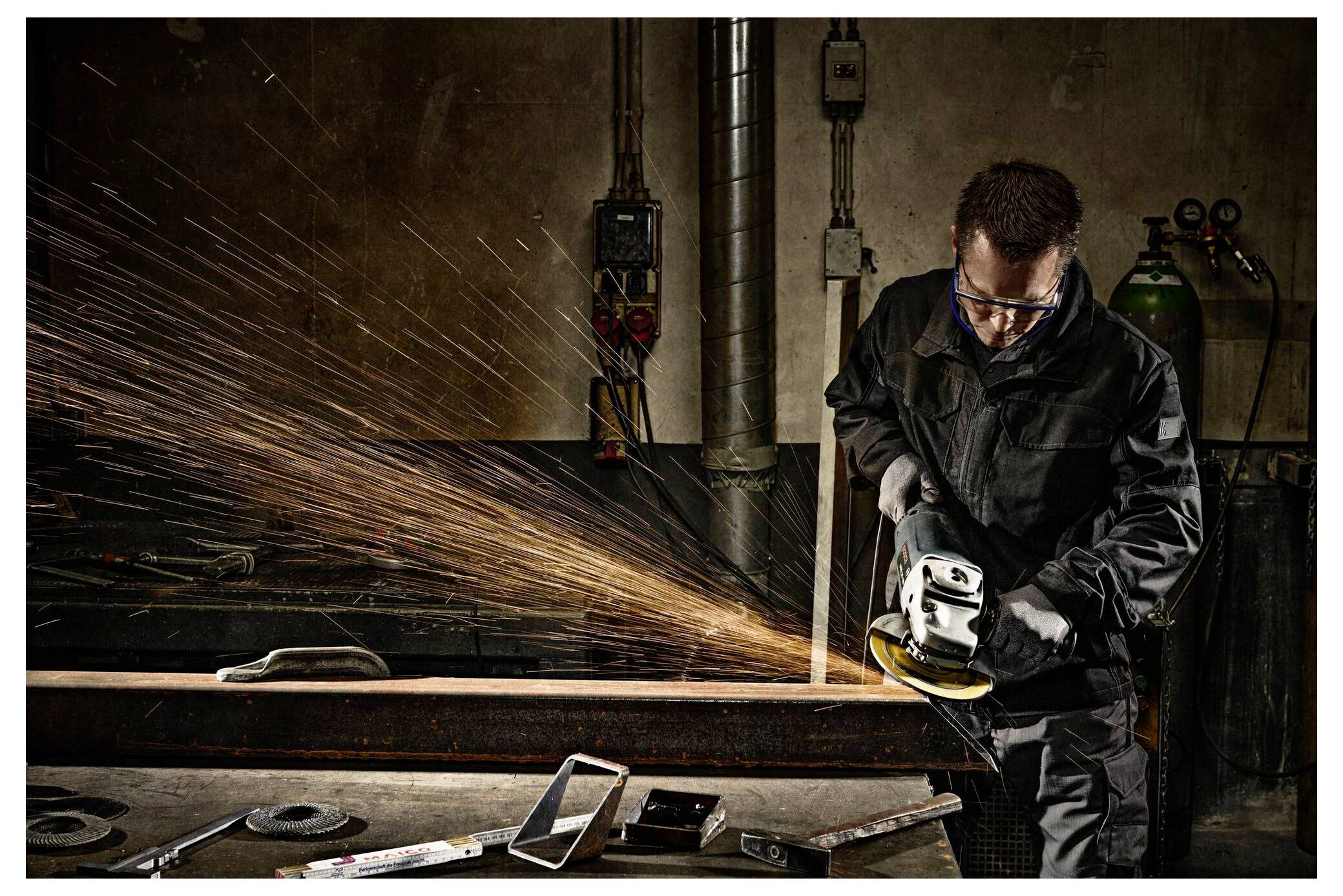 Eine Person in Schutzausrüstung bedient einen Handwinkelschleifer und erzeugt Funken, während sie an einem Metallträger in einer industriellen Werkstattsumgebung arbeitet.