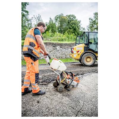 Ein Bauarbeiter in orangefarbener Sicherheitsausrüstung bedient eine Straßenschneidemaschine auf beschädigtem Pflaster, während ein Lader in der Nähe geparkt ist, umgeben von Grünpflanzen.