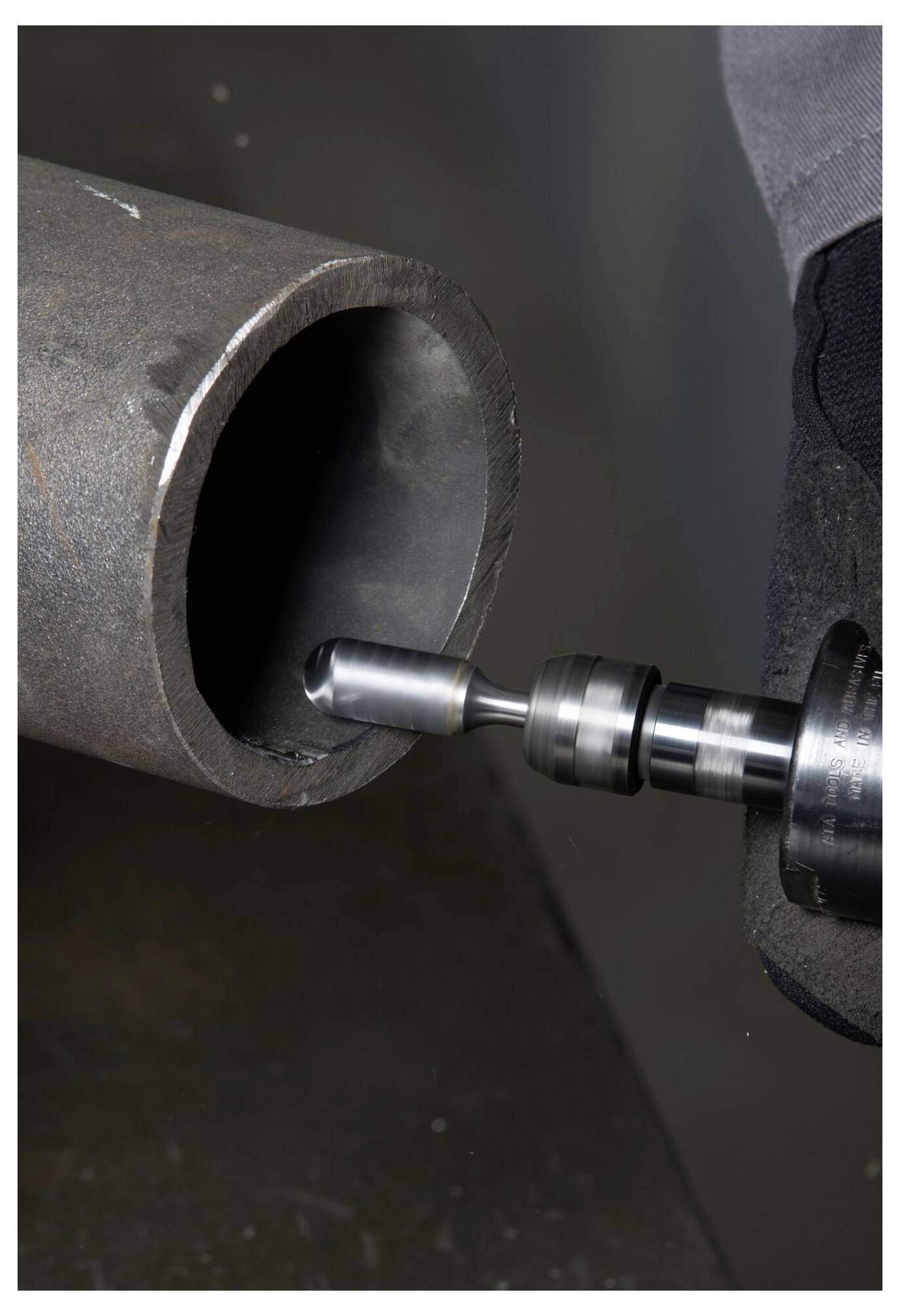 Eine Nahaufnahme einer Bohrmaschine in einer Handschuhen Hand, die in ein großes Metallrohr bohrt und eine mechanische oder industrielle Umgebung andeutet.