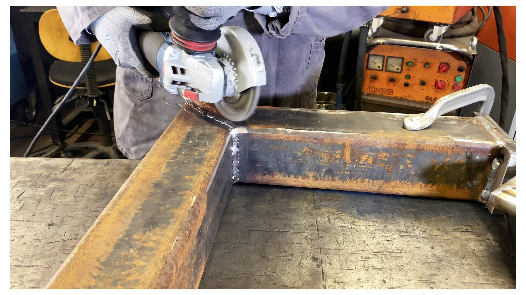 Eine Person trägt Handschuhe und verwendet einen Winkelschleifer, um eine Schweißnaht an einem großen, rostigen Metallrahmen in einer Werkstattumgebung zu glätten.