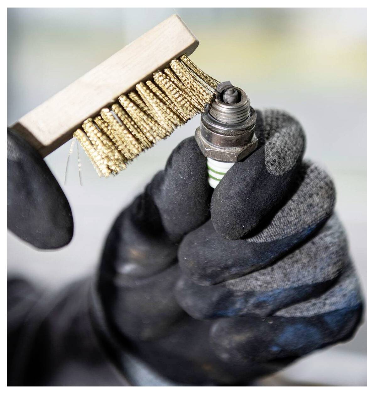 Eine Handschuh tragende Hand hält eine Zündkerze, während sie deren Spitze mit einer Drahtbürste reinigt und damit Wartungs- oder Reparaturarbeiten andeutet.