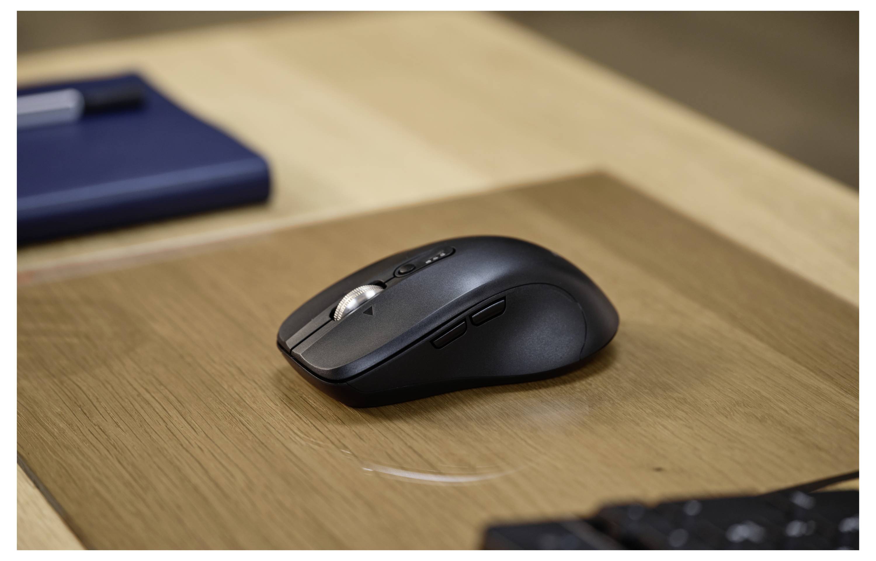 Eine schwarze Computer-Maus mit Seitenknöpfen liegt auf einer Holzoberfläche, wobei ein blaues Notizbuch und ein Stift im Hintergrund teilweise zu sehen sind.