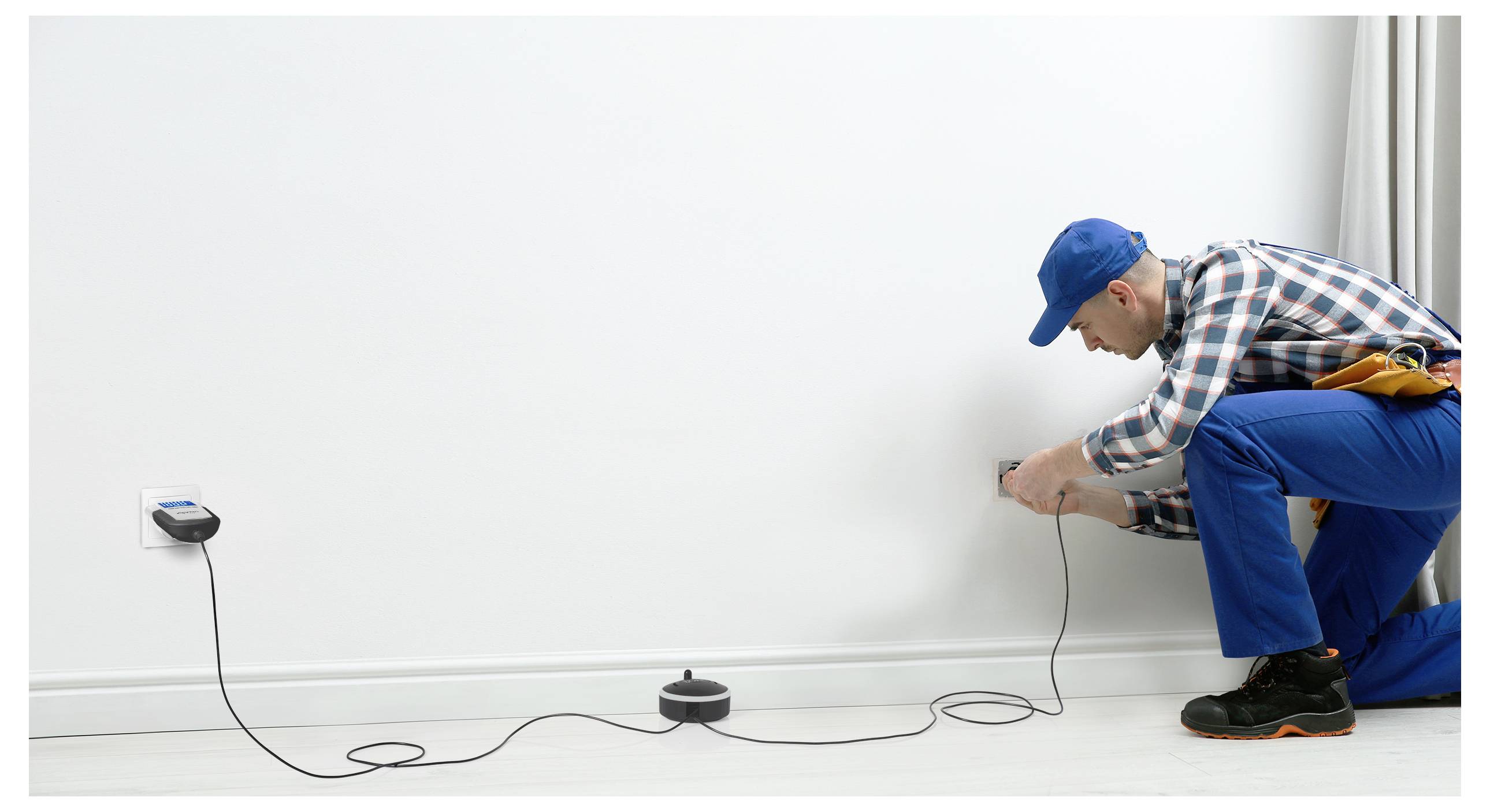 Elektriker in blauen Overalls und Mütze kniet neben einer Wandsteckdose und verlegt ein Stromkabel über den Boden in einem leeren Zimmer.