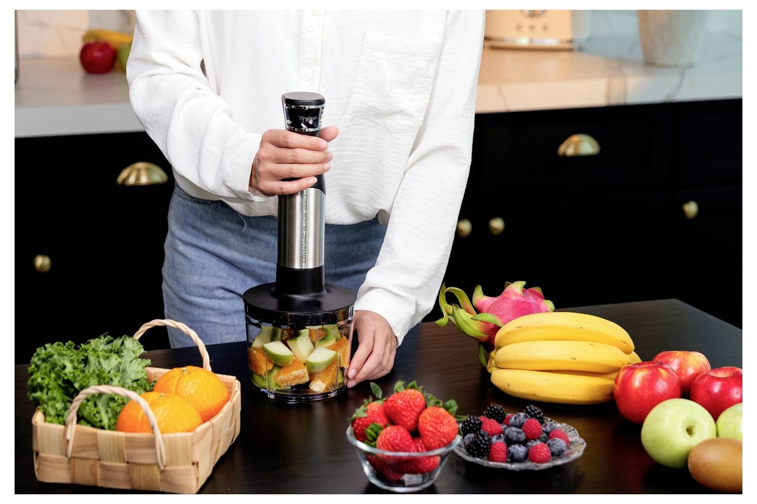 Eine Person benutzt einen tragbaren Küchenzerkleinerer auf einer Küchenarbeitsplatte, umgeben von Obst und Gemüse wie Bananen, Beeren und Grünblattgemüse.