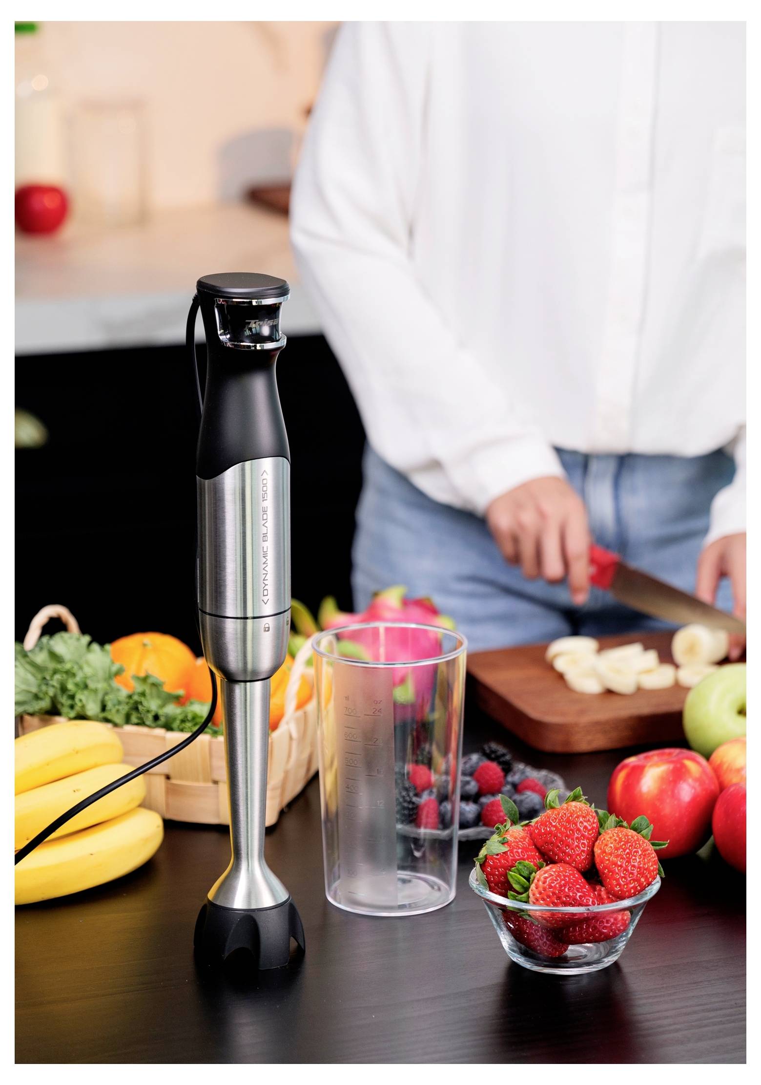 Eine Person schneidet Bananen in einer Küche neben einem Stabmixer, wobei verschiedene Früchte wie Erdbeeren, Bananen und Äpfel auf der Arbeitsplatte liegen.