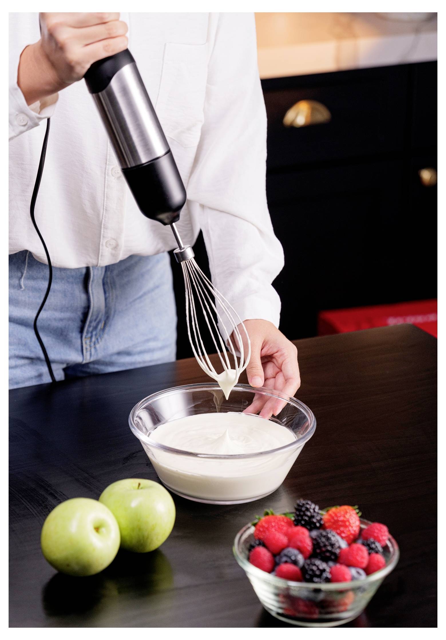 Eine Person verwendet einen Handrührer, um Sahne in einer Schüssel auf einem Küchentisch zu schlagen, wobei Äpfel und eine Schüssel mit Beeren in der Nähe stehen.