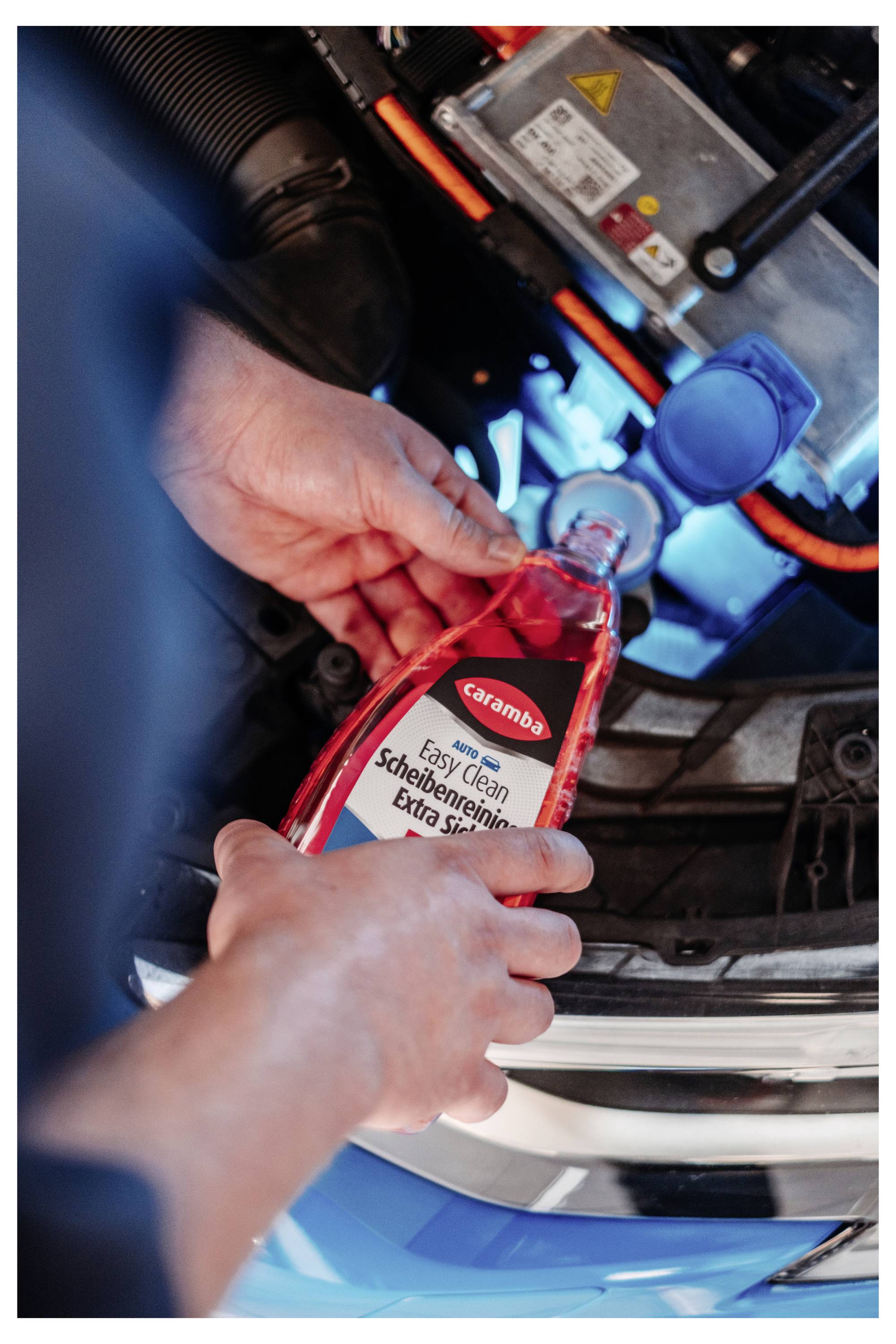 Hände gießen roten „Easy Clean Scheibenenteiser" Scheibenentfroster in einen Automotorraum neben einer blauen Kappe.
