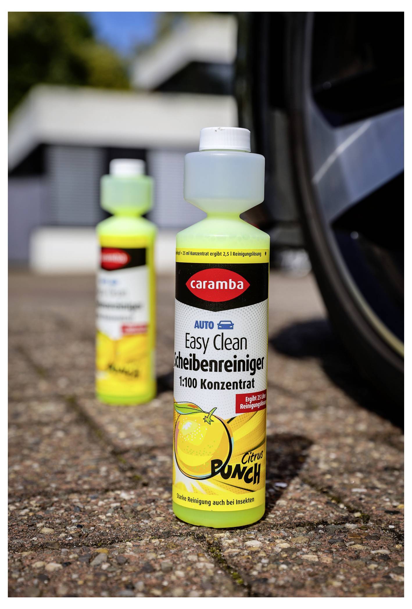 Ein 'Caramba Easy Clean Scheibenreiniger' Zitrusfrische Glasreiniger-Flasche im Fokus, mit einer weiteren, unscharfen Flasche im Hintergrund auf Pflastersteinen.