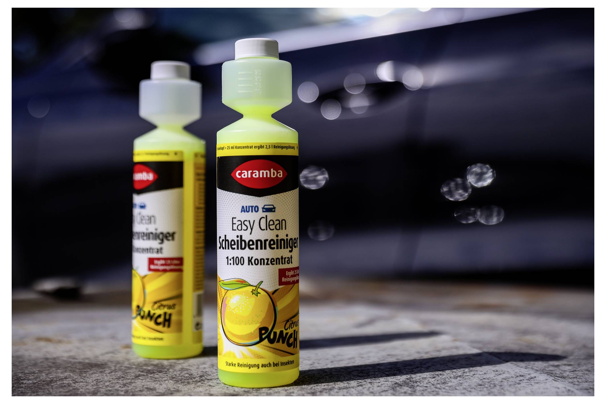 Zwei Flaschen 'Caramba Auto Easy Clean Scheibenreiniger', ein zitronenduftender Scheibenwaschmittel, stehen auf dem Pflaster. Im Hintergrund ein unscharfes Auto.