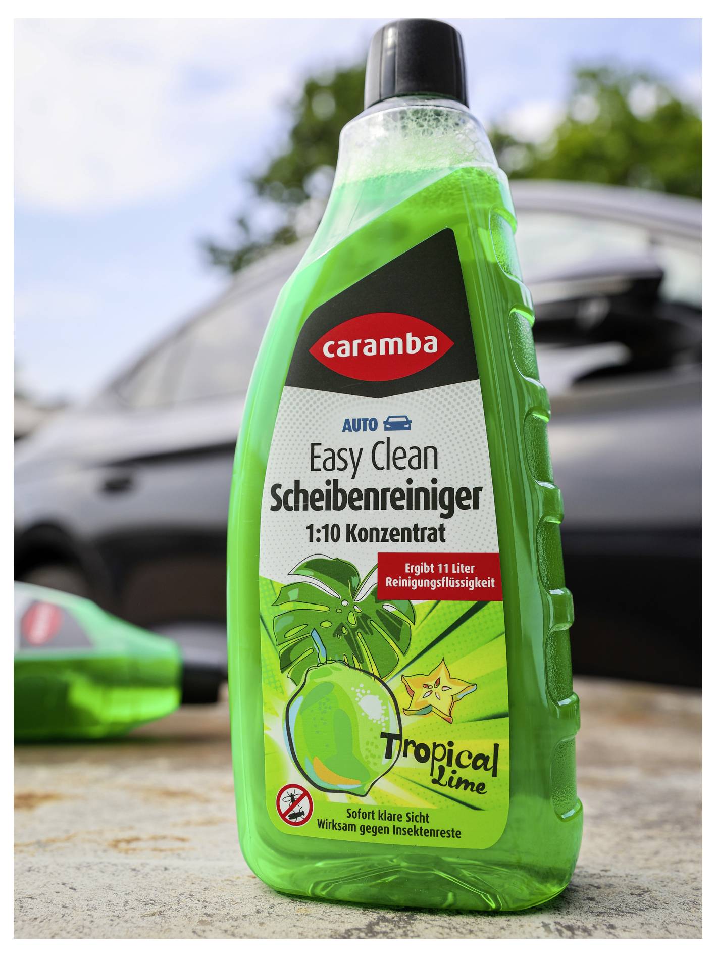 Eine grüne Flasche 'Caramba Easy Clean' Glasreiniger mit tropischer Limetten-Duftnote, vor einem verschwommenen Hintergrund mit einem Auto und Bäumen.