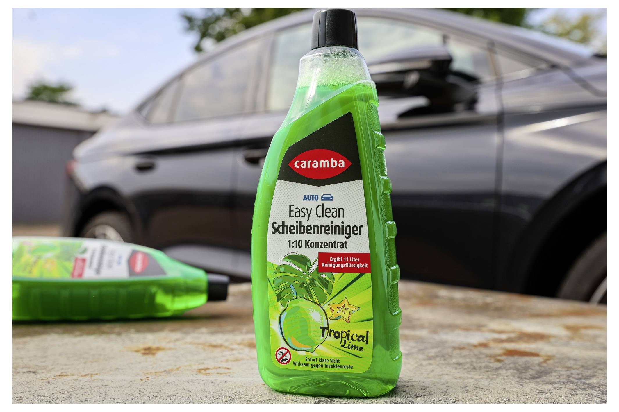 Eine grüne Flasche 'Easy Clean Scheibenreiniger' Autoreiniger auf einer Betonoberfläche, mit einem unscharfen Auto im Hintergrund.