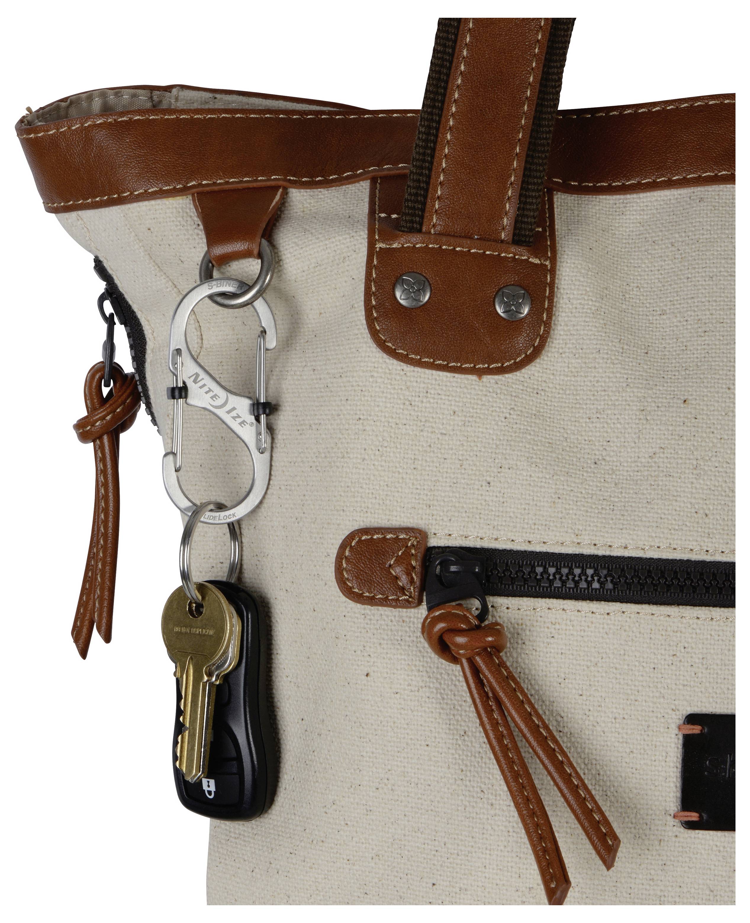 Eine beige Leinwandtasche mit braunen Lederriemen hat einen kleinen Karabiner befestigt, an dem Metallschlüssel und ein Schlüsselanhänger hängen.