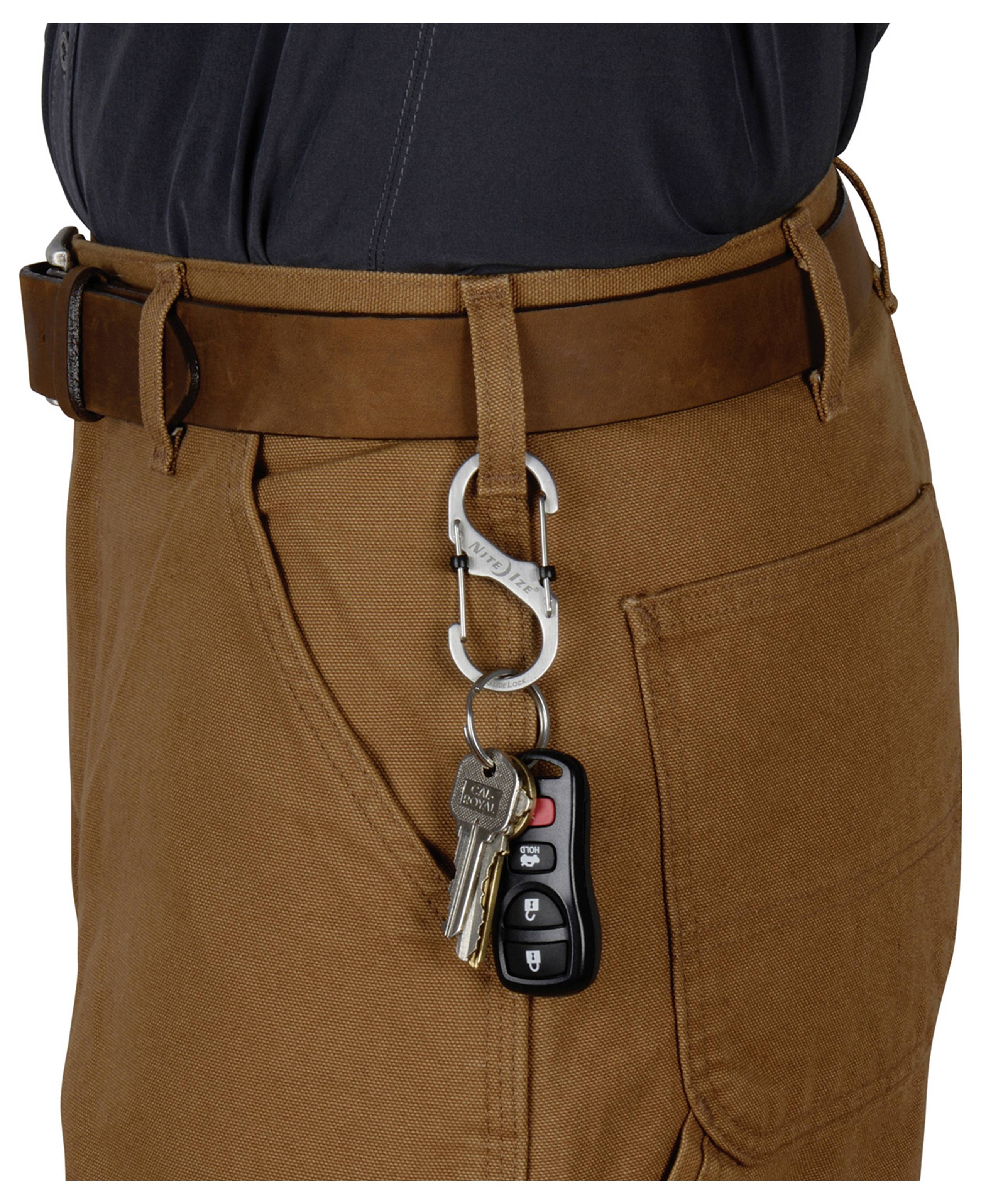 Ein Metallkarabiner, der an einem braunen Gürtel befestigt ist, hält einen Satz Schlüssel und eine Autoferndedienung, aufgenommen vor einer braunen Hose.