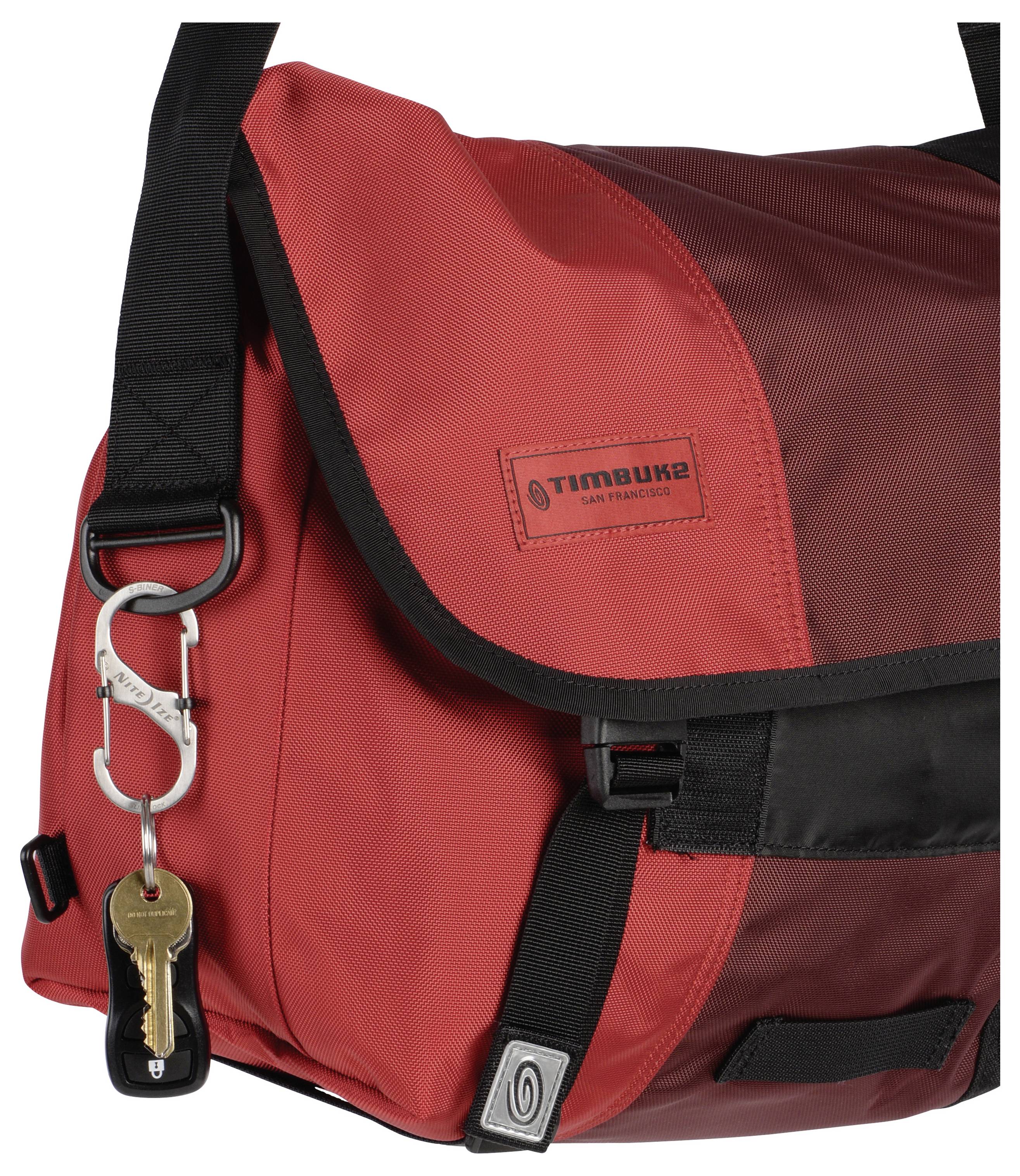 Eine rote Umhängetasche mit einem schwarzen Trageriemen, die einen kleinen Metallhaken mit einem Schlüssel trägt. Die Tasche trägt ein Logo mit der Aufschrift "timbuk2 San Francisco".
