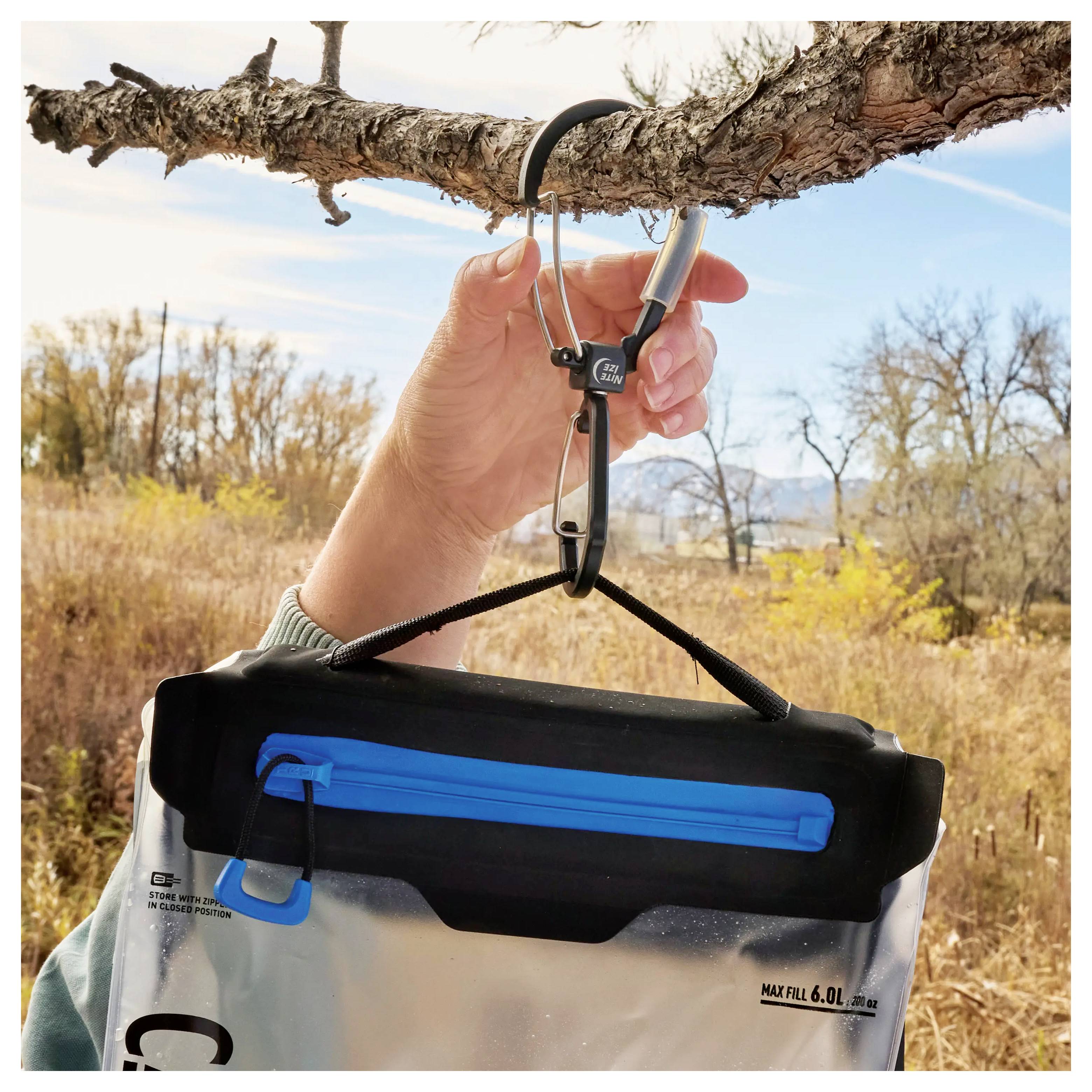Eine Hand hängt einen Wasserbeutel mit einem Karabiner von einem Baumast ab, vor der Kulisse von Herbstbäumen und Gras in einer natürlichen Umgebung.