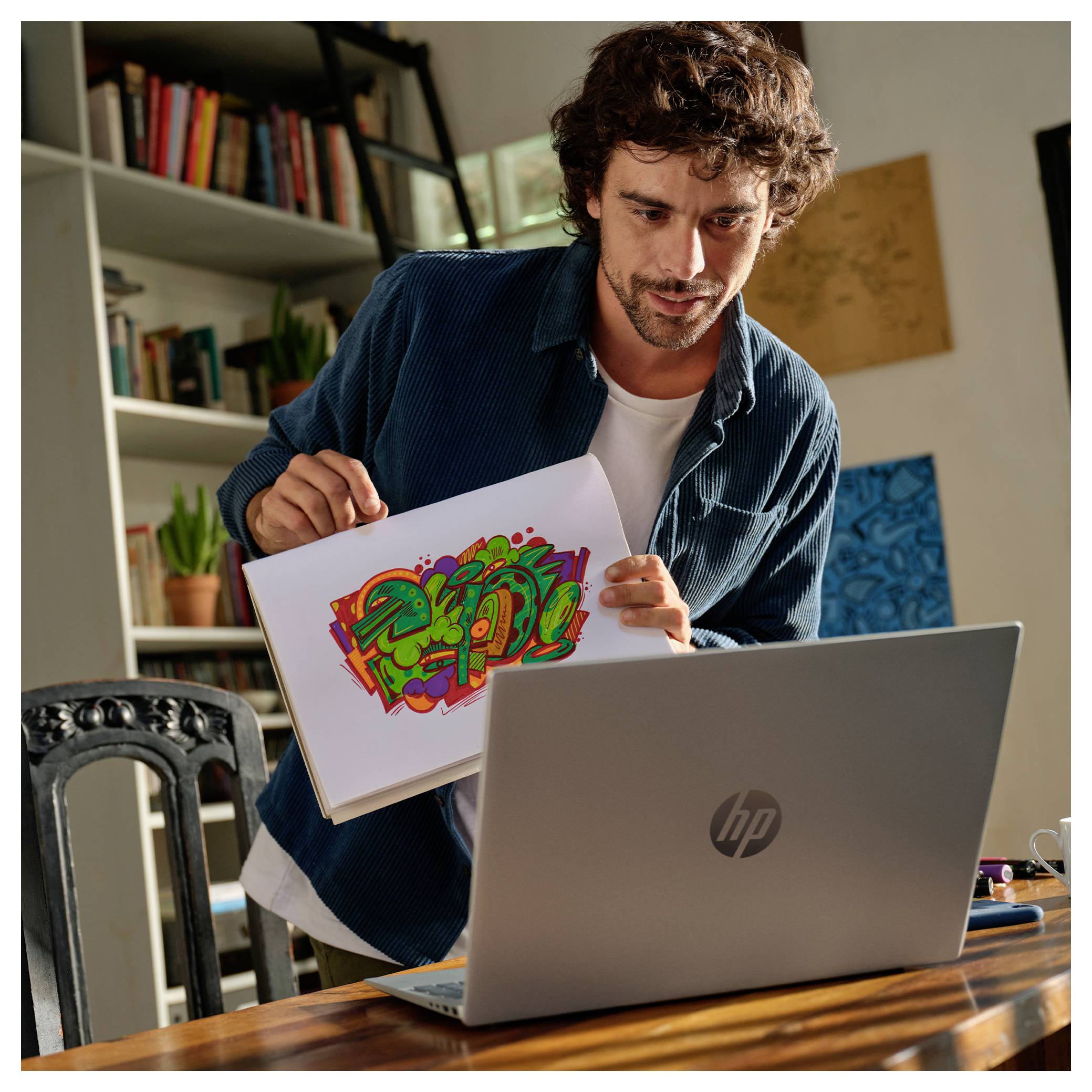 Eine Person zeigt einen bunten Graffiti-Entwurf in eine Laptop-Webcam und präsentiert offensichtlich Kunst in einem gemütlichen, mit Büchern gefüllten Raum.