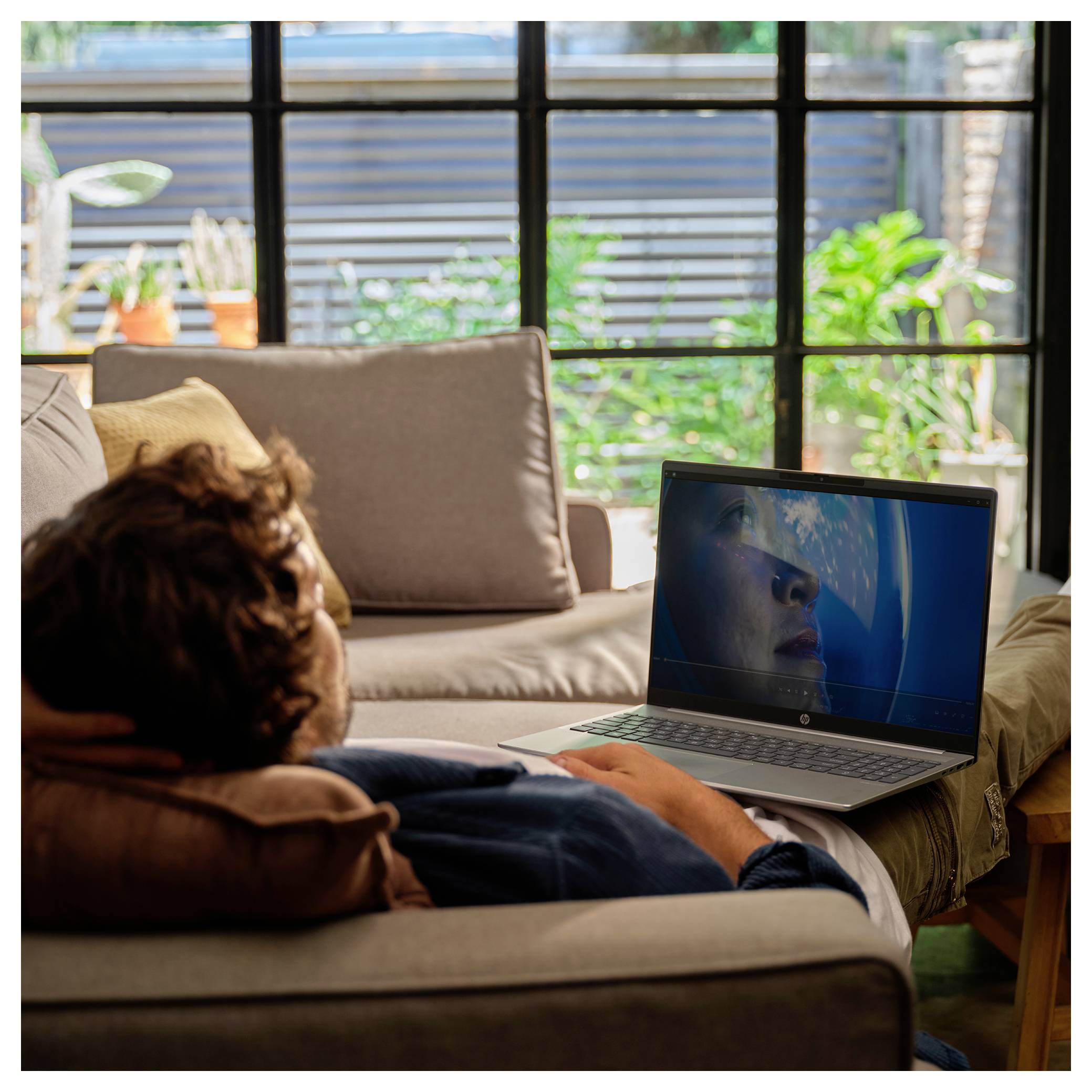 Eine Person liegt auf einem Sofa in einem gemütlichen Raum und schaut auf einen Laptop-Bildschirm, der eine Nahaufnahme eines Gesichts neben einem Globus zeigt, während draußen Pflanzen zu sehen sind.