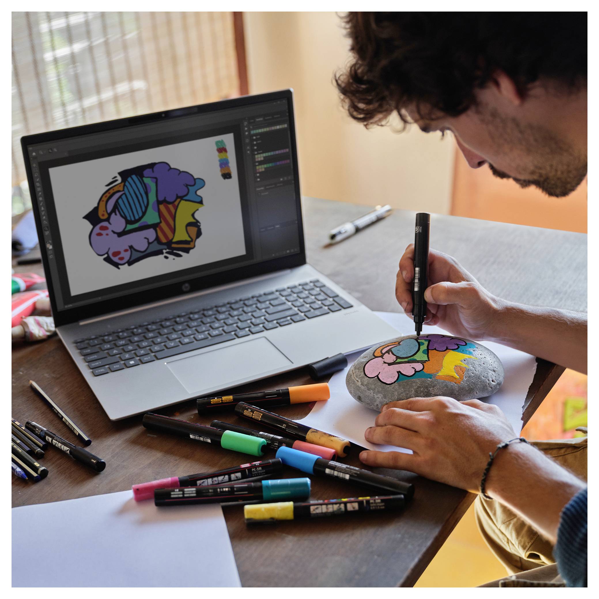 Eine Person malt einen bunten Cartoon-Charakter auf einen Stein und verwendet dabei eine digitale Kunstvorlage von einem Laptop. Farbmarker sind auf dem Tisch verstreut.