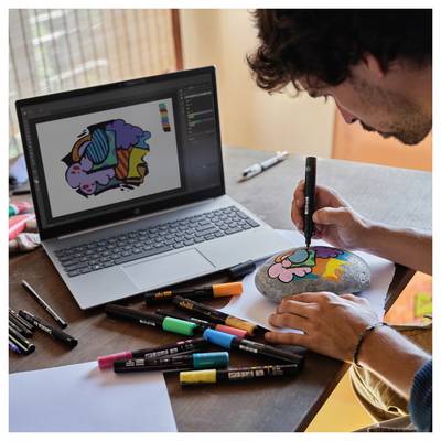 Eine Person malt einen bunten Cartoon-Charakter auf einen Stein und verwendet dabei eine digitale Kunstvorlage von einem Laptop. Farbmarker sind auf dem Tisch verstreut.