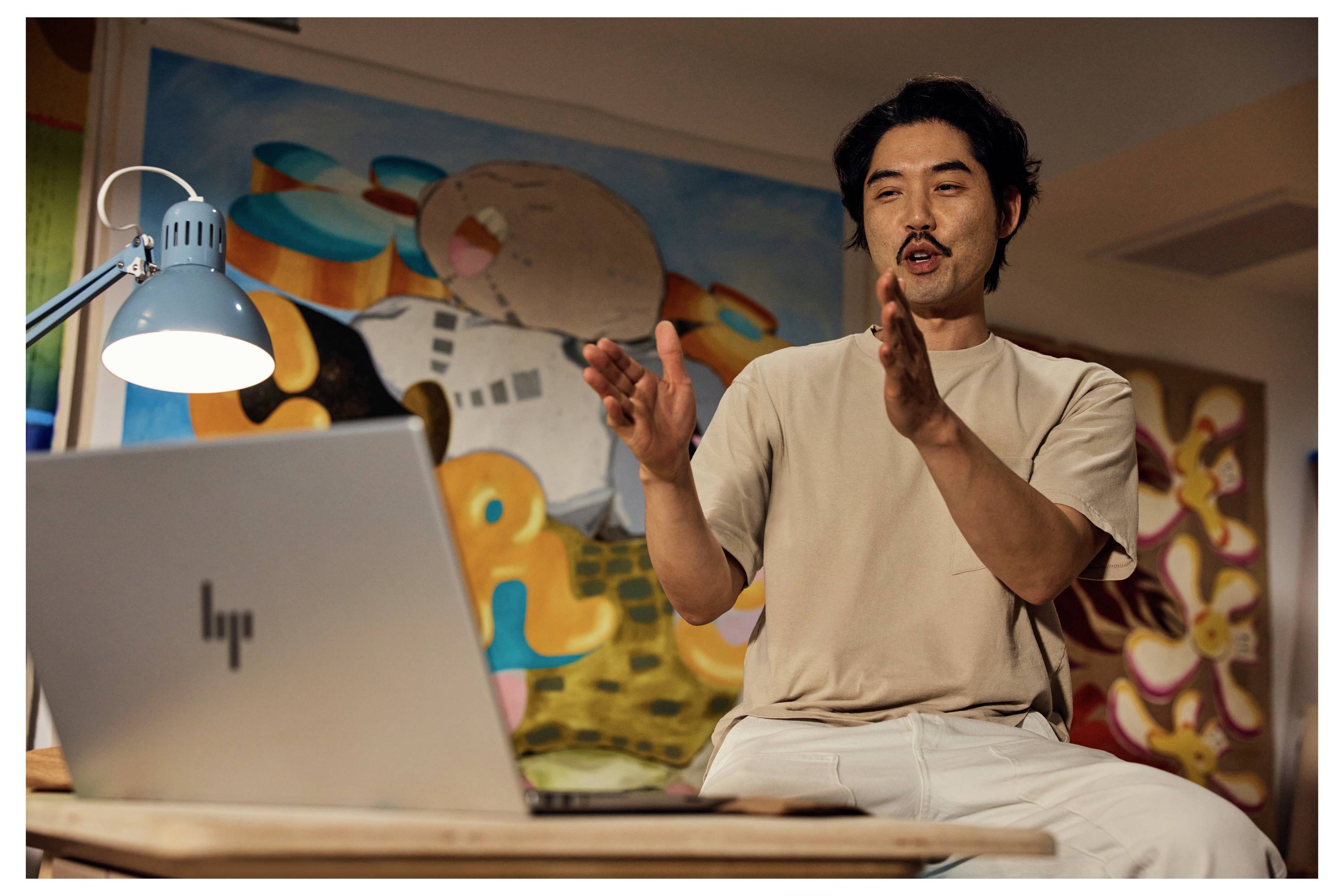 Eine Person gestikuliert während sie vor einem Laptop spricht, in einem bunten Raum mit abstrakten Kunstwerken im Hintergrund.