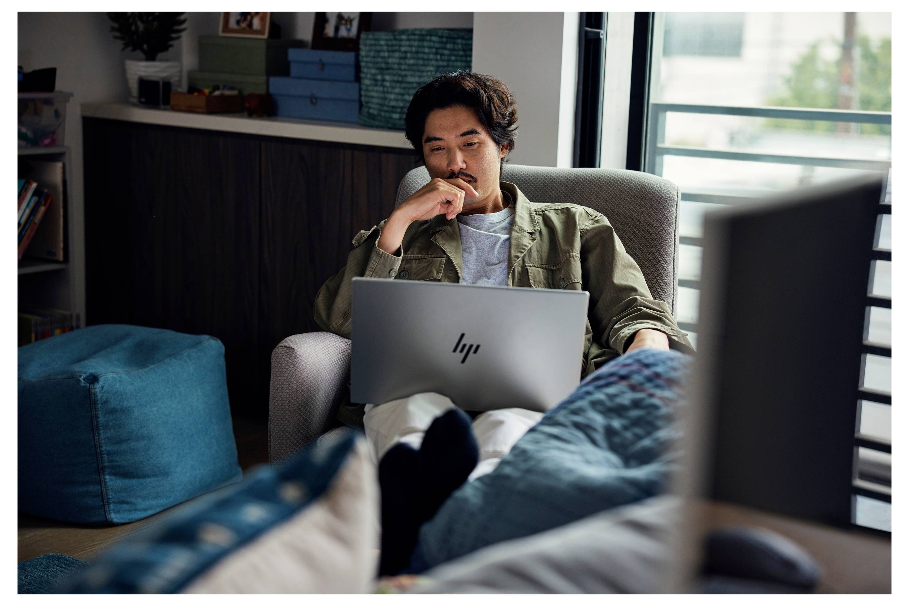 Eine Person sitzt zu Hause auf einem Stuhl und arbeitet an einem Laptop. Der Raum hat eine entspannte, lockere Atmosphäre mit petrolfarbener Möbel und natürlichem Licht.