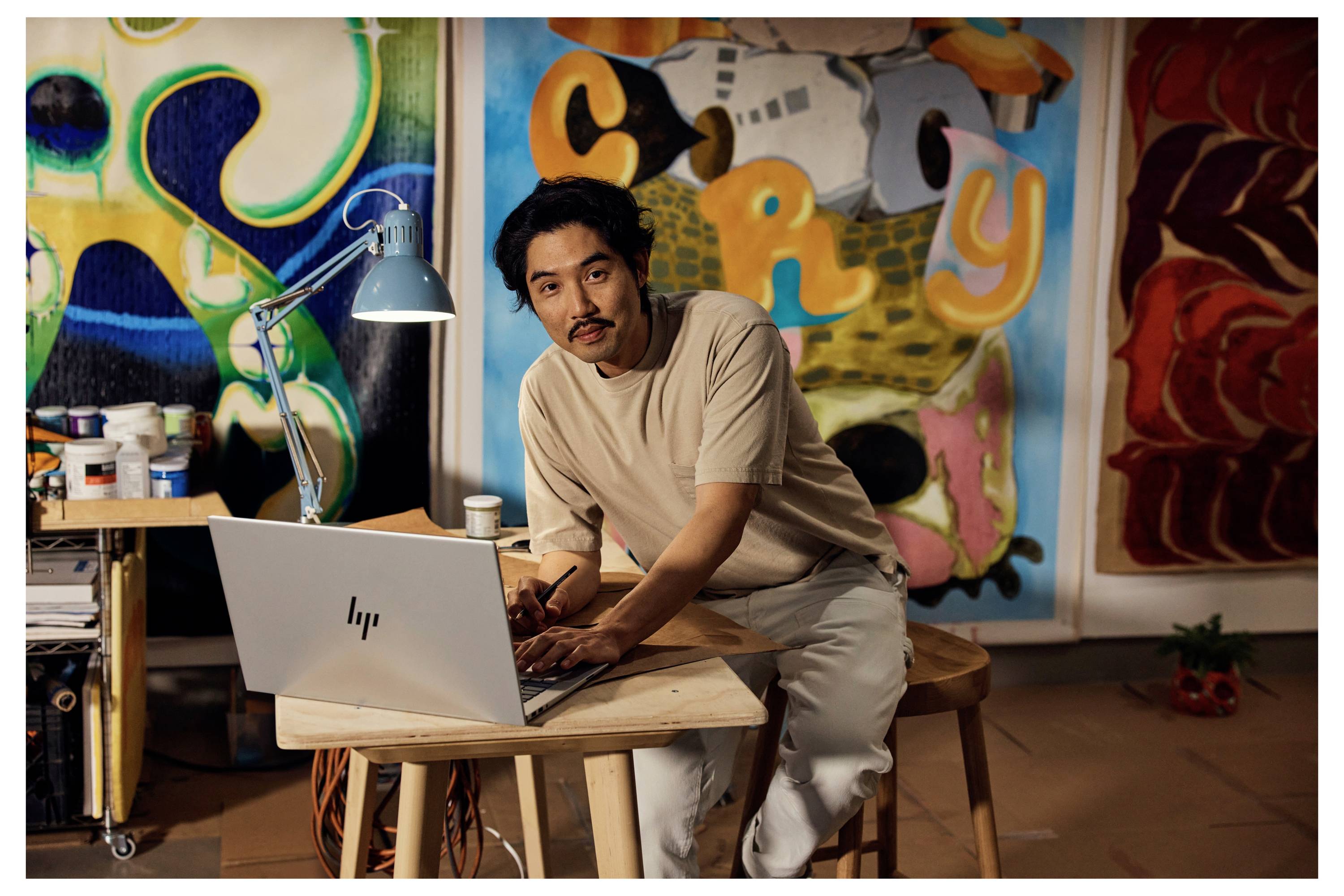 Ein Künstler, der an einem Laptop in einem hell erleuchteten Studio arbeitet, umgeben von farbenfrohen abstrakten Gemälden und Kunstutensilien.