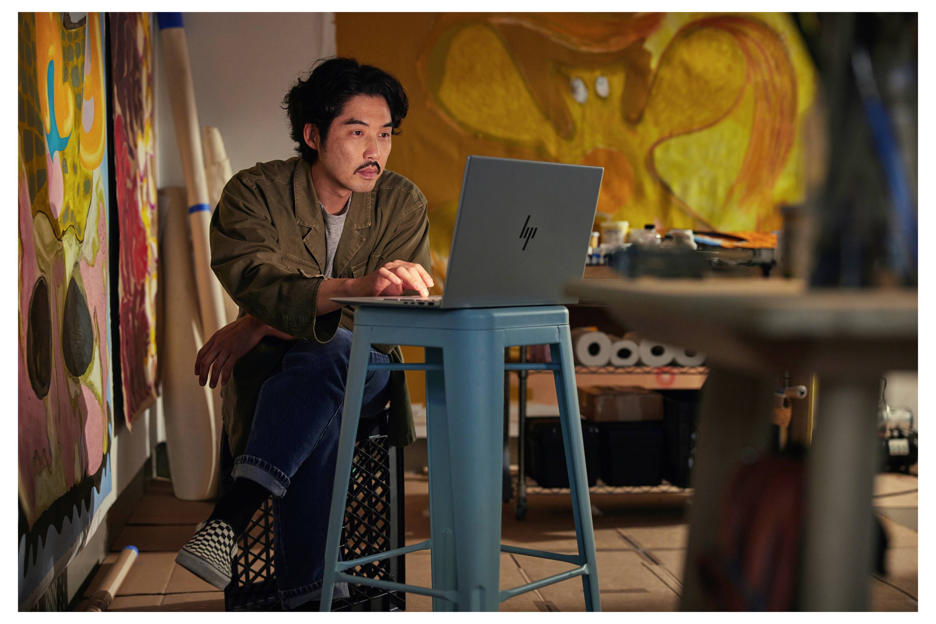 Eine Person sitzt an einem blauen Tisch und arbeitet an einem Laptop in einem Kunstatelier, umgeben von Gemälden und Künstlerbedarf.