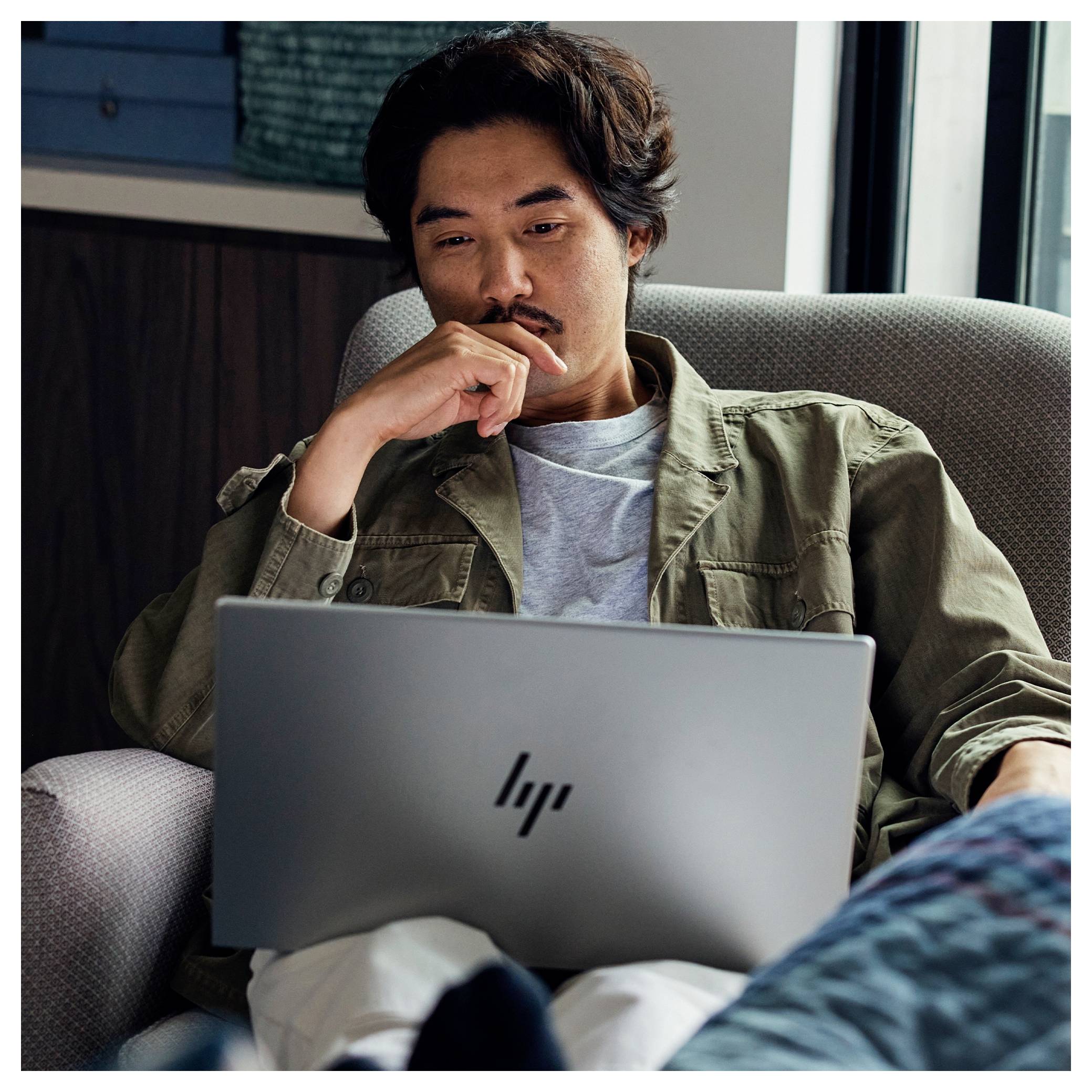 „Ein Mann sitzt auf einem Stuhl, nachdenklich blickend, während er an einem Laptop arbeitet, was Fokus und Konzentration vermittelt."