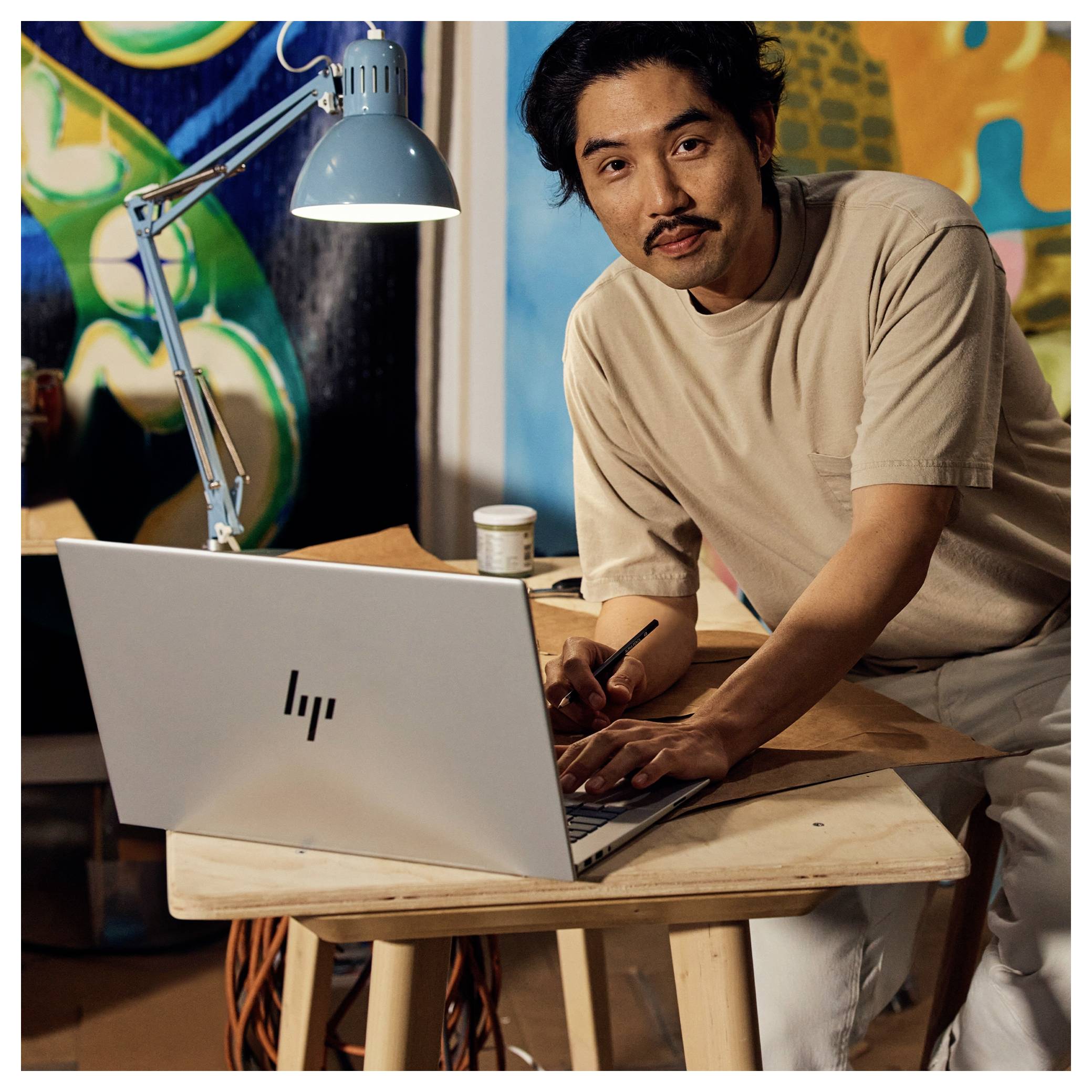 Ein Mann in einem lässigen T-Shirt sitzt an einem Schreibtisch mit einem Laptop und blickt in die Kamera. Im Hintergrund befindet sich ein buntes Kunstwerk und eine blaue Schreibtischlampe.