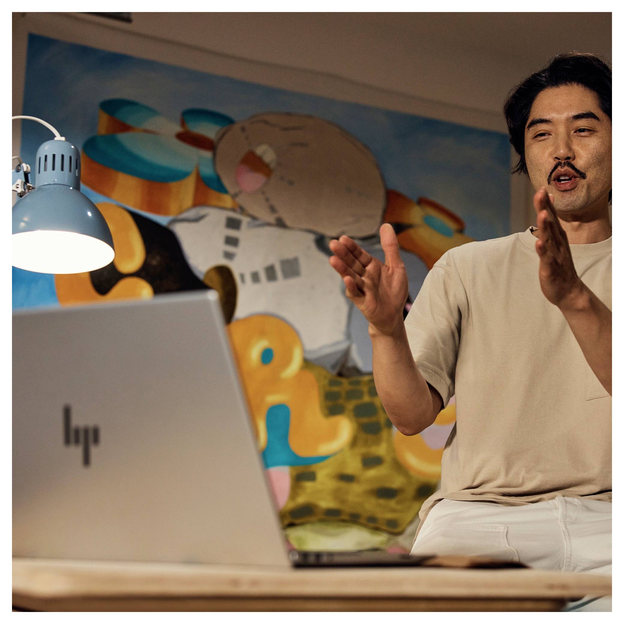 Eine Person gestikuliert aufgeregt, während sie an einem Tisch mit einem Laptop sitzt, in einem hell gestalteten Raum mit einem gemalten Wandgemälde im Hintergrund.