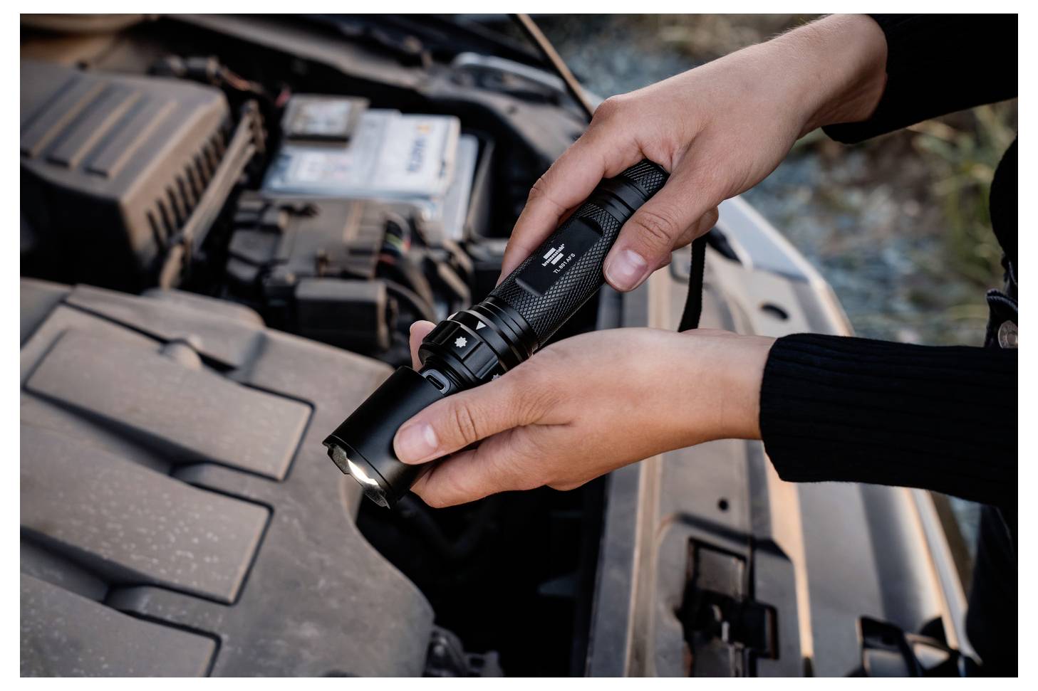 Eine Person hält eine Taschenlampe, um einen geöffneten Automotor zu untersuchen, was auf Wartungs- oder Reparaturarbeiten hindeutet.