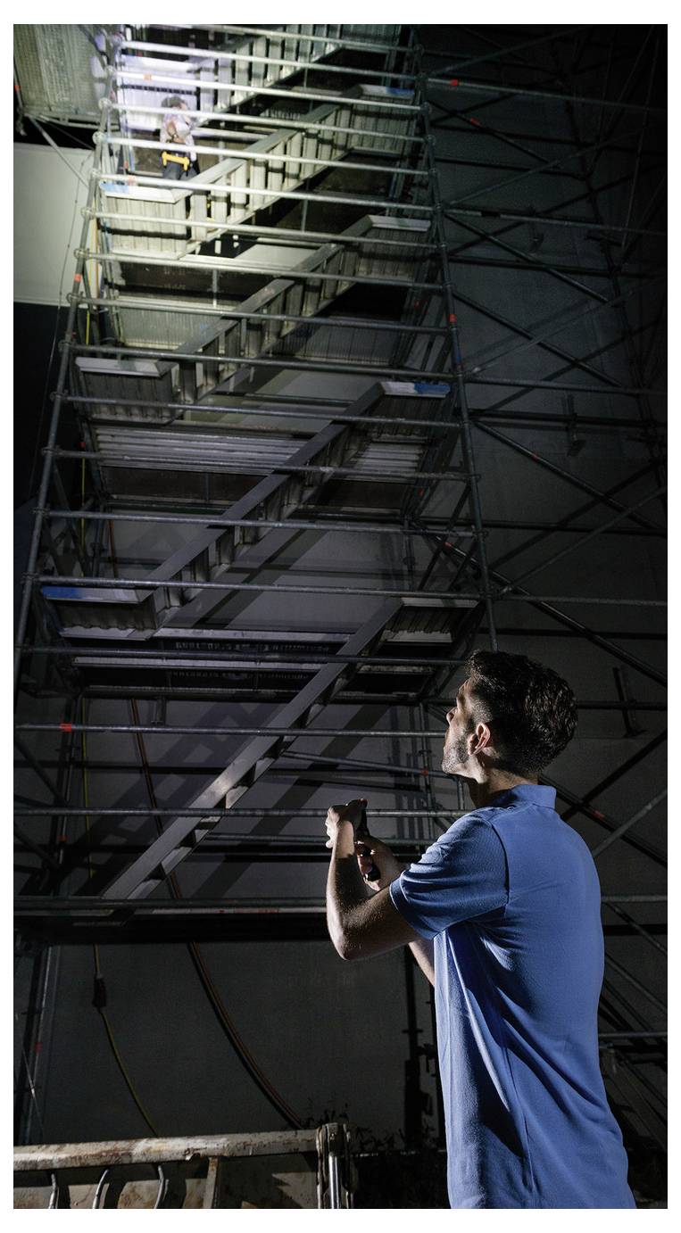 Eine Person in einem blauen Hemd blickt mit einer Taschenlampe zu einer hohen, industriellen Treppe hinauf. Eine weitere Gestalt ist teilweise an der Spitze sichtbar.