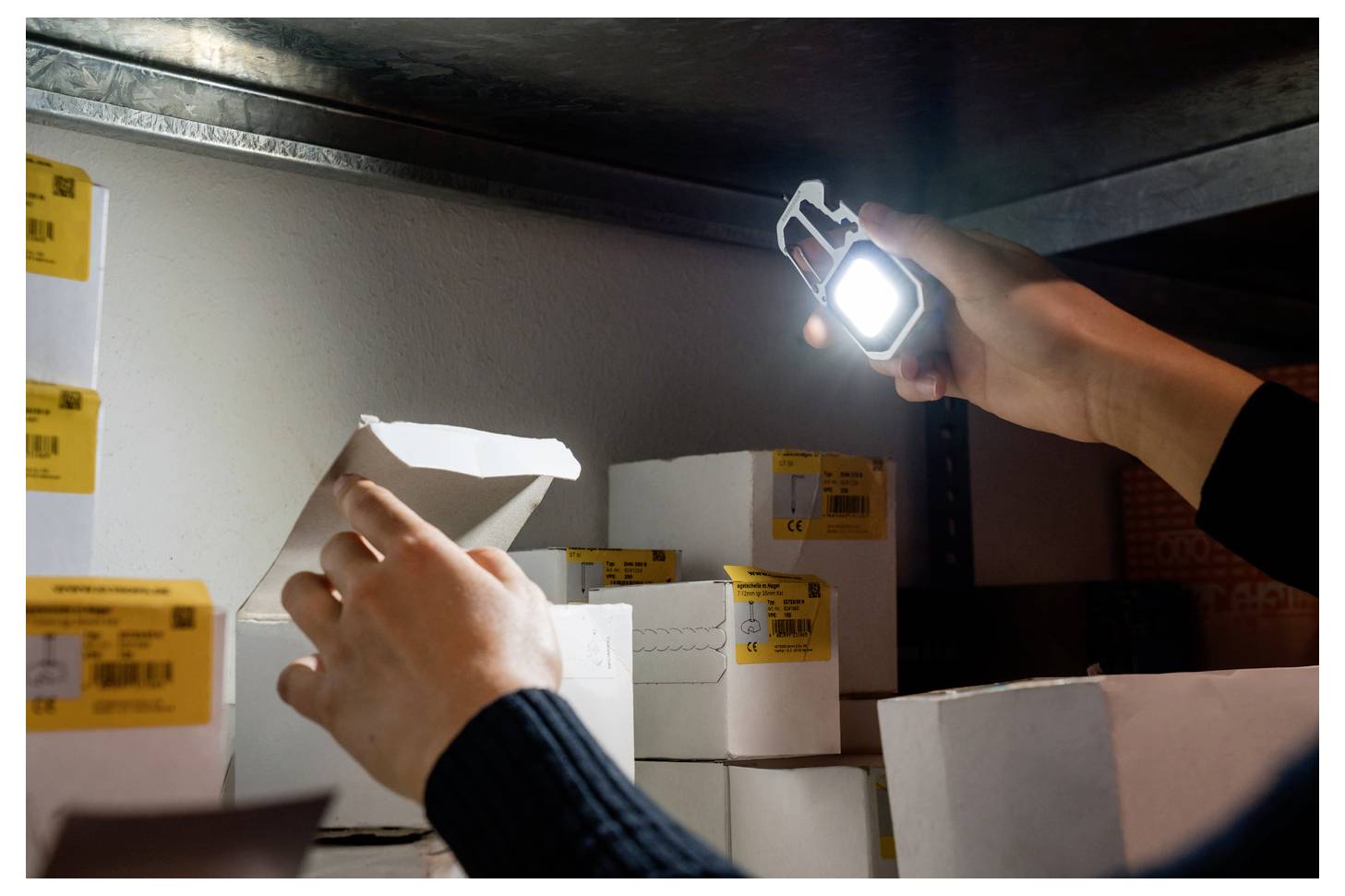 Eine Person, die eine Taschenlampe hält, öffnet ein Paket auf einem Regal in einem dunkel beleuchteten Lagerbereich und untersucht Kartons mit gelben Etiketten.