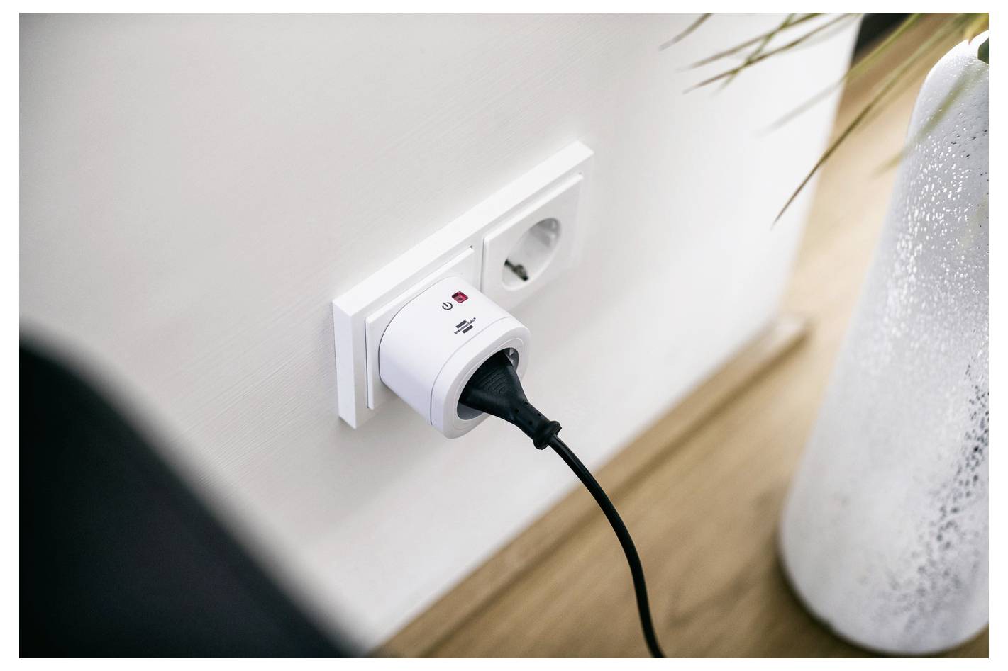 Eine weiße Wandsteckdose mit einem schwarzen Netzstecker, der eingesteckt ist. Das Gerät zeigt eine Spannungsanzeige und rote und grüne Kontrollleuchten.