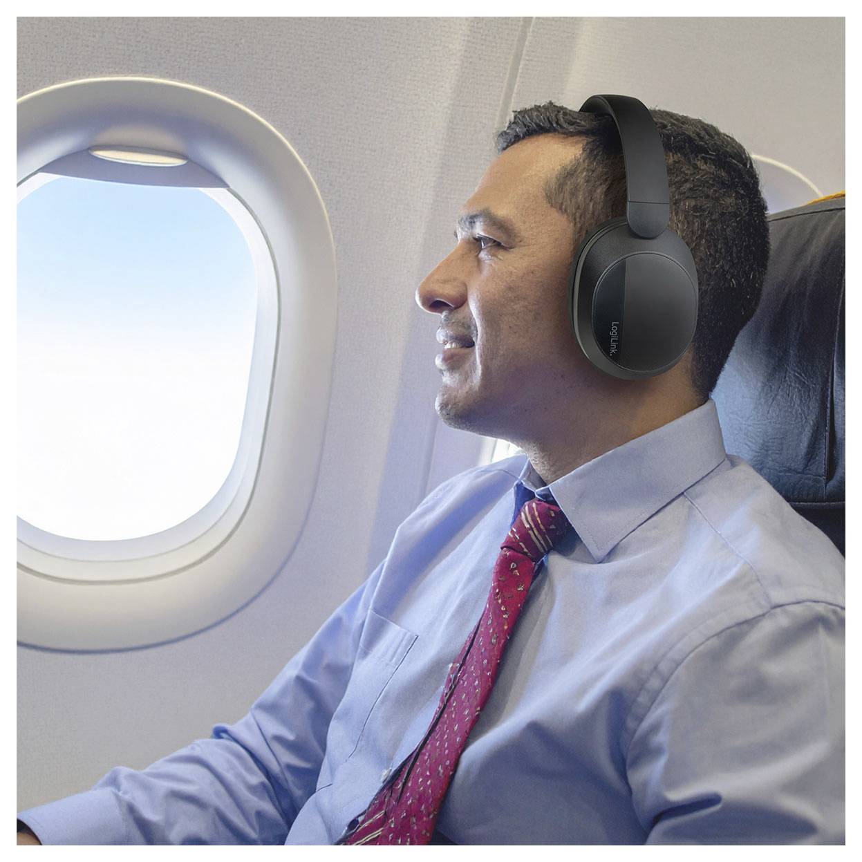 Eine Person mit Kopfhörern, die in einem Flugzeug sitzt, blickt aus dem Fenster in einen klaren Himmel. Sie trägt ein Hemd und eine Krawatte.