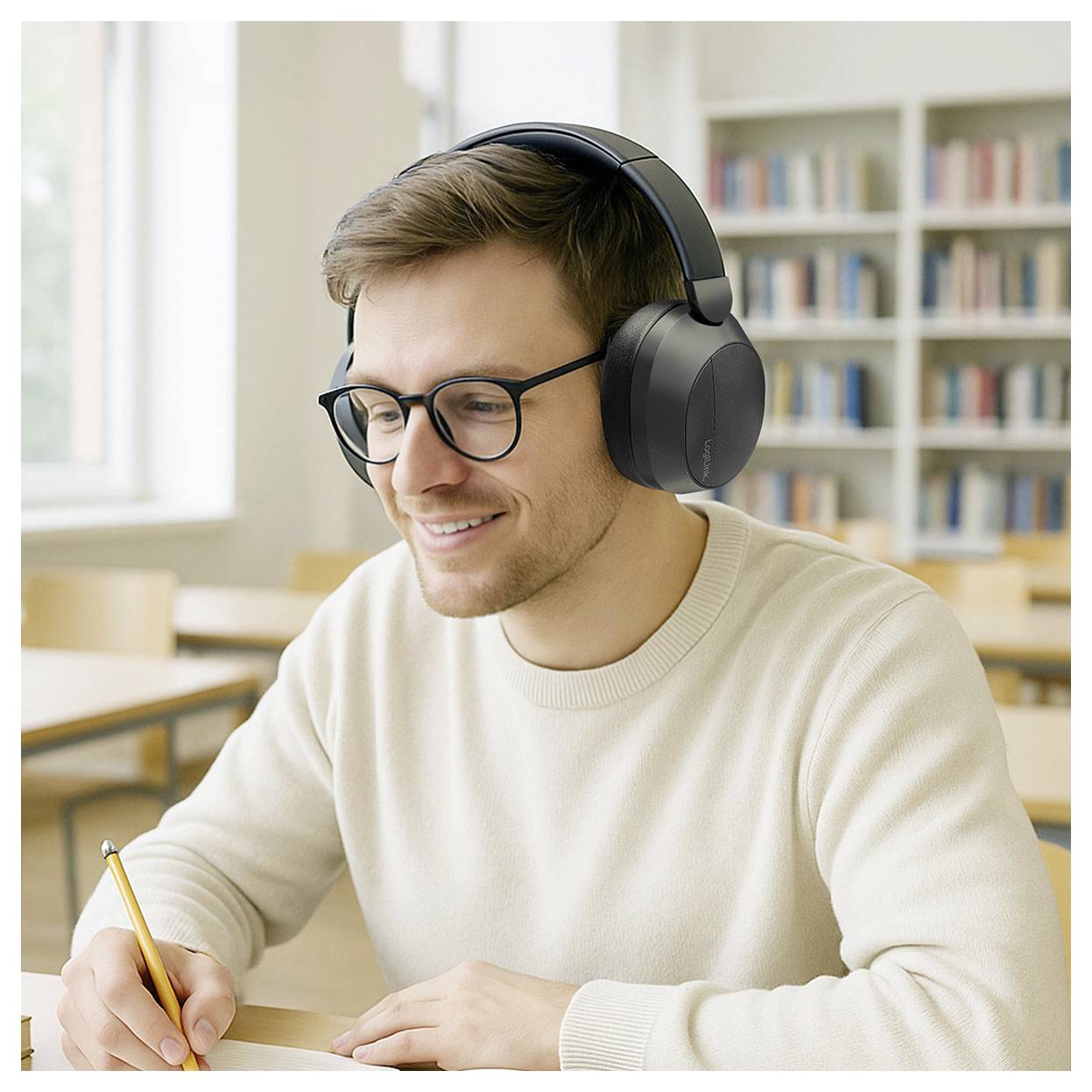 Eine Person mit Kopfhörern und Brille lächelt, während sie in einem Notizbuch in einer Bibliotheksumgebung schreibt. Im Hintergrund sind Bücherregale zu sehen.