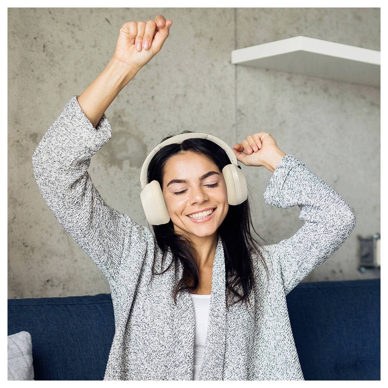 Eine Frau mit Kopfhörern lächelt, die Augen geschlossen, und hebt freudig ihre Arme, während sie offensichtlich Musik genießt. Sie sitzt auf einem Sofa in einem gemütlichen Raum.
