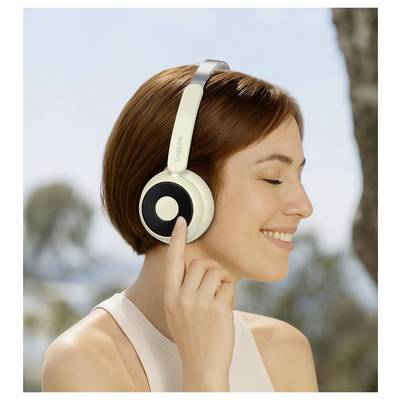 Eine lächelnde Person mit kurzen braunen Haaren trägt Over-Ear-Kopfhörer im Freien und genießt Musik oder Klänge, mit Bäumen und blauem Himmel im Hintergrund.