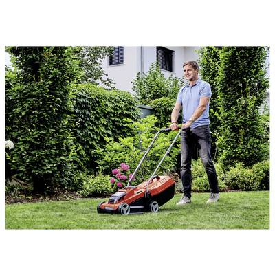 Ein Mann schiebt einen Rasenmäher auf einem grasbewachsenen Rasen. Er ist im Freien, umgeben von Grünflächen und einem blühenden Strauch, mit einem Haus im Hintergrund.