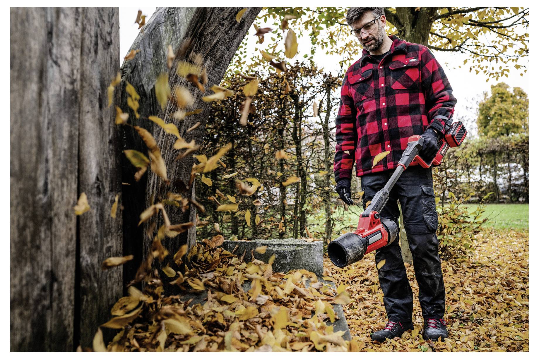 Ein Mann in einem roten Karohemd benutzt einen Laubbläser, um gefallene Blätter in einem Außenbereich mit Bäumen und einer Hecke im Hintergrund zu beseitigen.