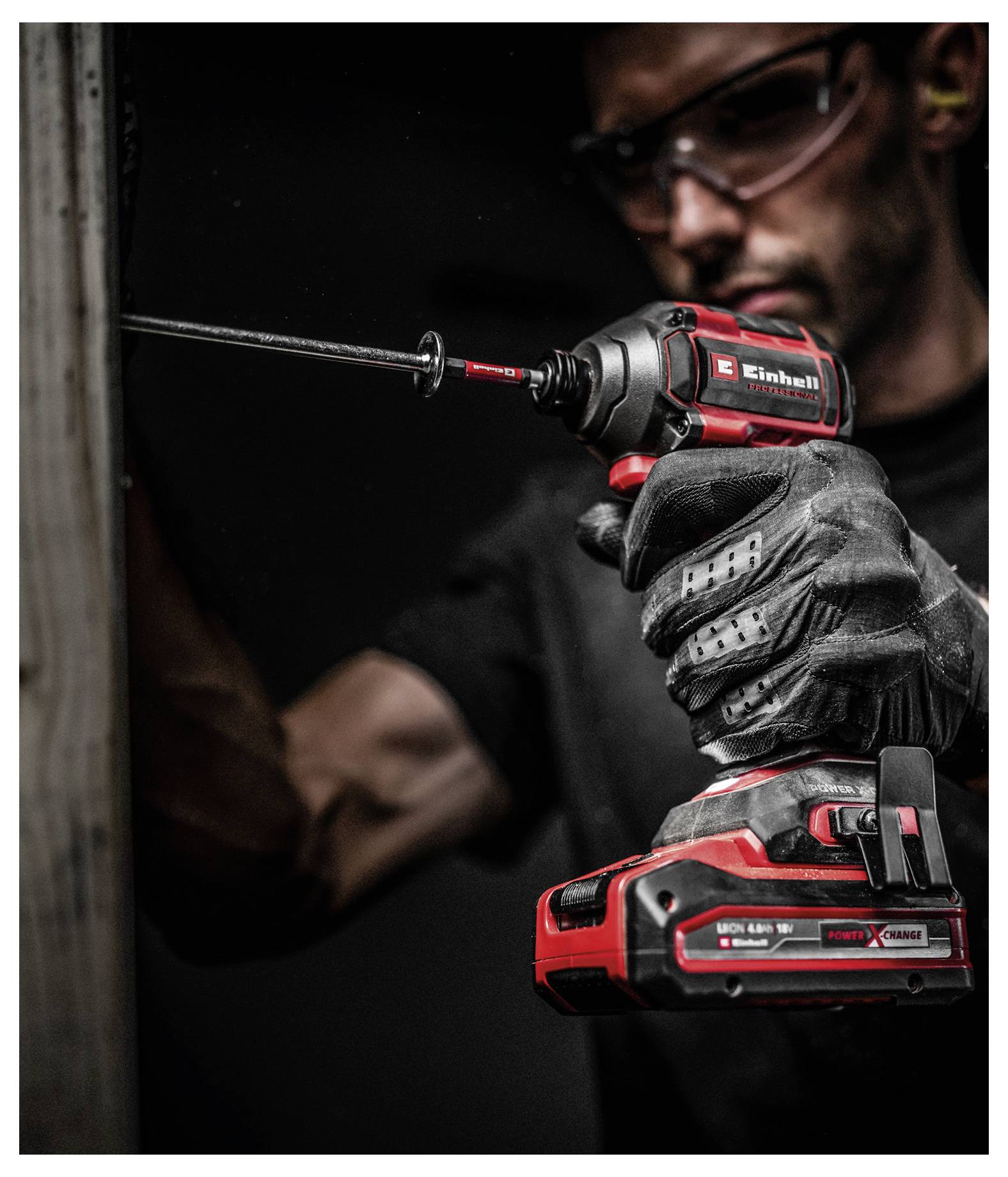 Eine Person mit Sicherheitsbrille und Handschuhen benutzt eine rote und schwarze elektrische Bohrmaschine auf einer Holzoberfläche und konzentriert sich auf ihre Aufgabe.