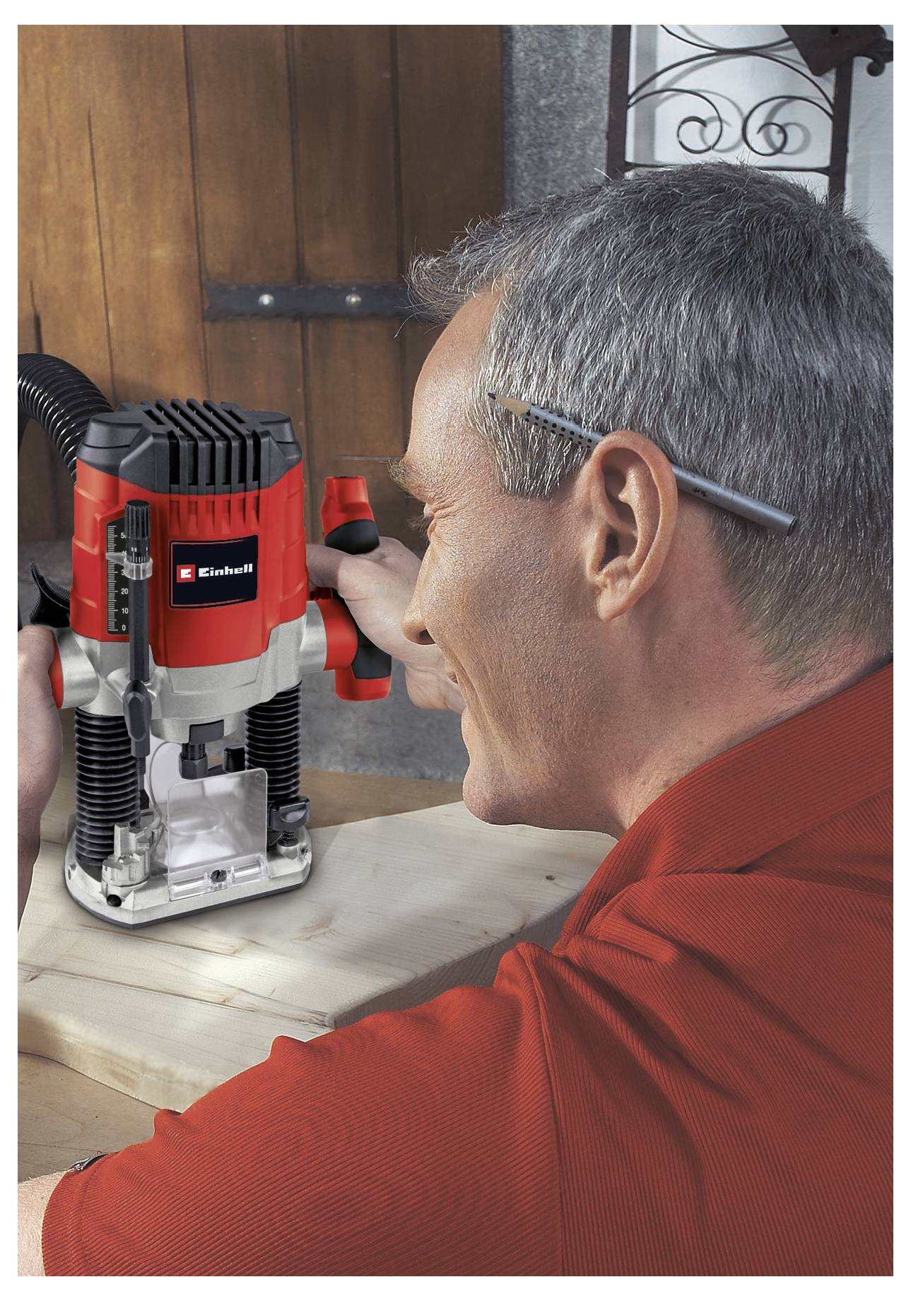 Eine Person, die einen roten elektrischen Router auf Holz verwendet, konzentriert auf ihre Aufgabe, mit einem Bleistift hinter dem Ohr, in einer Werkstattumgebung.