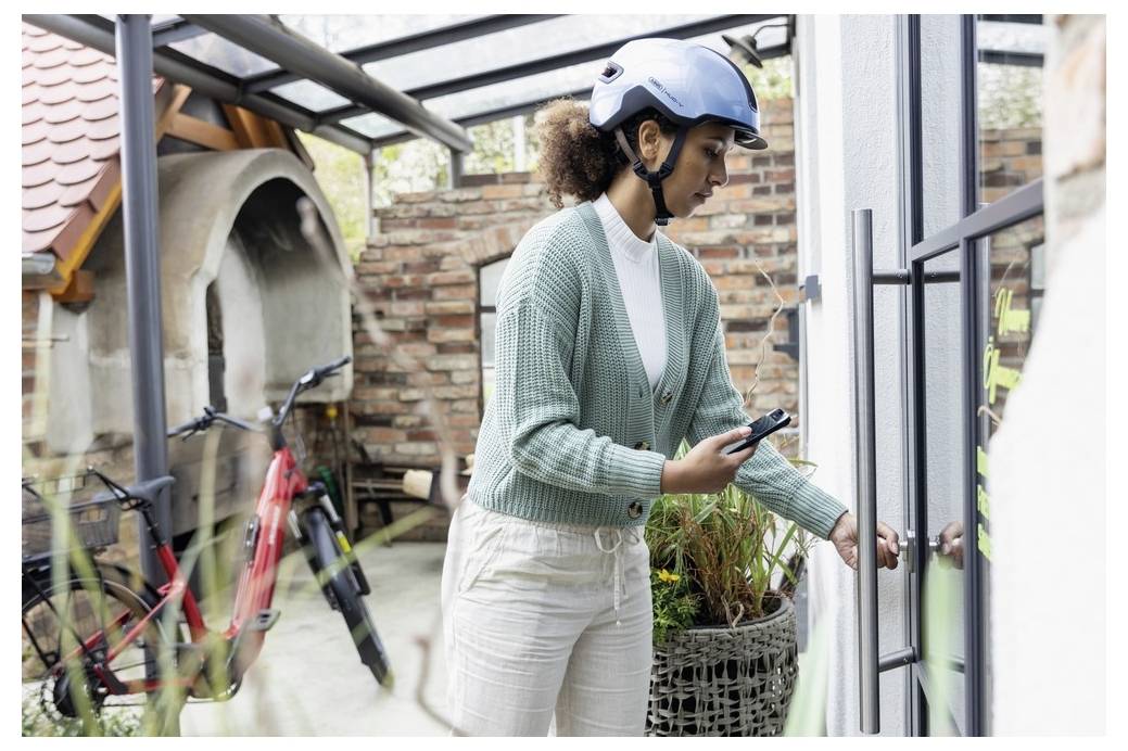 Eine Person mit einem blauen Helm öffnet eine Glastür, in der Nähe eines roten Fahrrads im Freien. Die Umgebung scheint residential mit Ziegelwänden zu sein.
