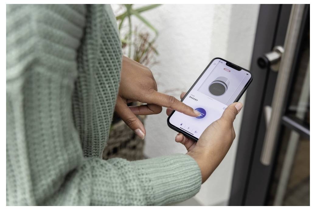 Eine Person in einem grünen Pullover nutzt eine Smartphone-App, um ein Smart-Türschloss zu entriegeln und präsentiert damit moderne Technologie und Sicherheit.