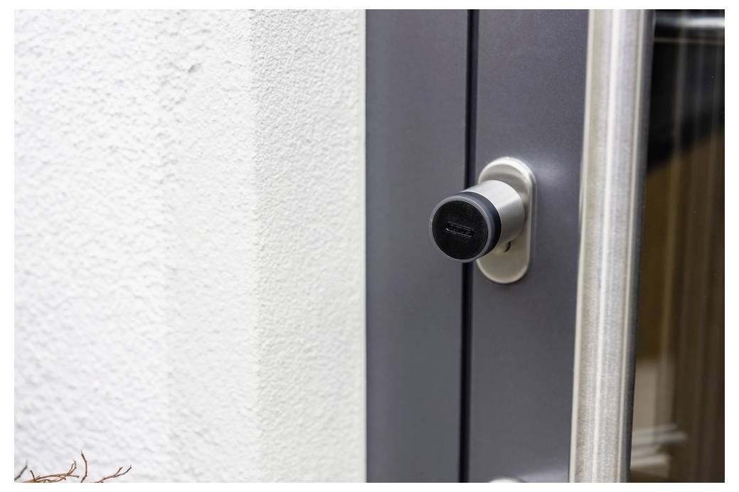 Eine silberne Tür mit einem schwarzen Smart-Lock installiert, neben einer weißen strukturierten Wand, was moderne Sicherheit und Design andeutet.