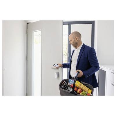 Eine Person in einem Anzug hält ein Telefon, um eine Tür zu entriegeln, während sie einen Korb mit Lebensmitteln trägt und damit die Nutzung einer Smart-Lock-Technologie demonstriert.