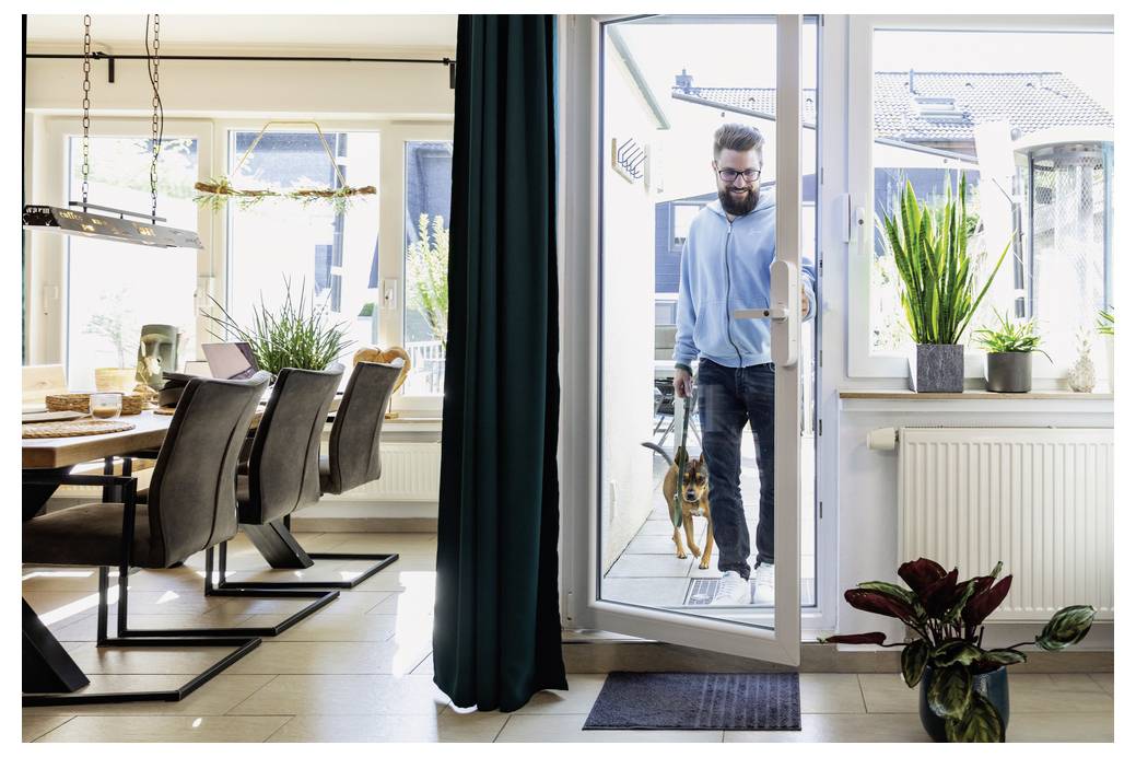 Ein Mann mit Bart betritt durch eine Glastür einen hell erleuchteten Speiseraum und hält einen kleinen Hund an der Leine. Der Raum hat Stühle, einen Tisch und Topfpflanzen in der Nähe der Fenster.