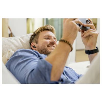 Ein Mann mit Kopfhörern und einem lässigen Shirt lächelt, während er entspannt auf einem Sofa liegt und auf ein Smartphone schaut, und wirkt gelassen und zufrieden.