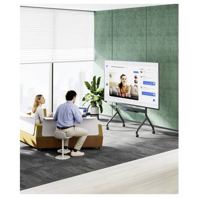 Zwei Personen in einem modernen Büro arbeiten zusammen, indem sie Laptops nutzen und einen großen Bildschirm verwenden, der eine Videokonferenz mit mehreren Teilnehmern zeigt.